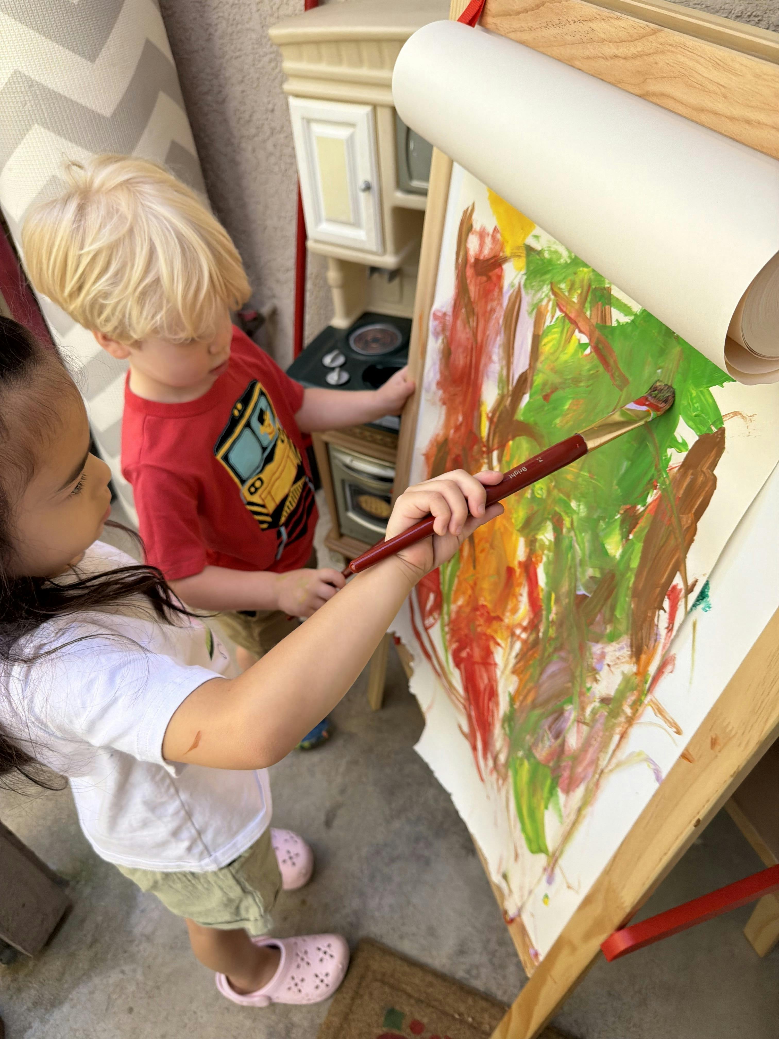 Child painting on an easel while another child stands next to the side
