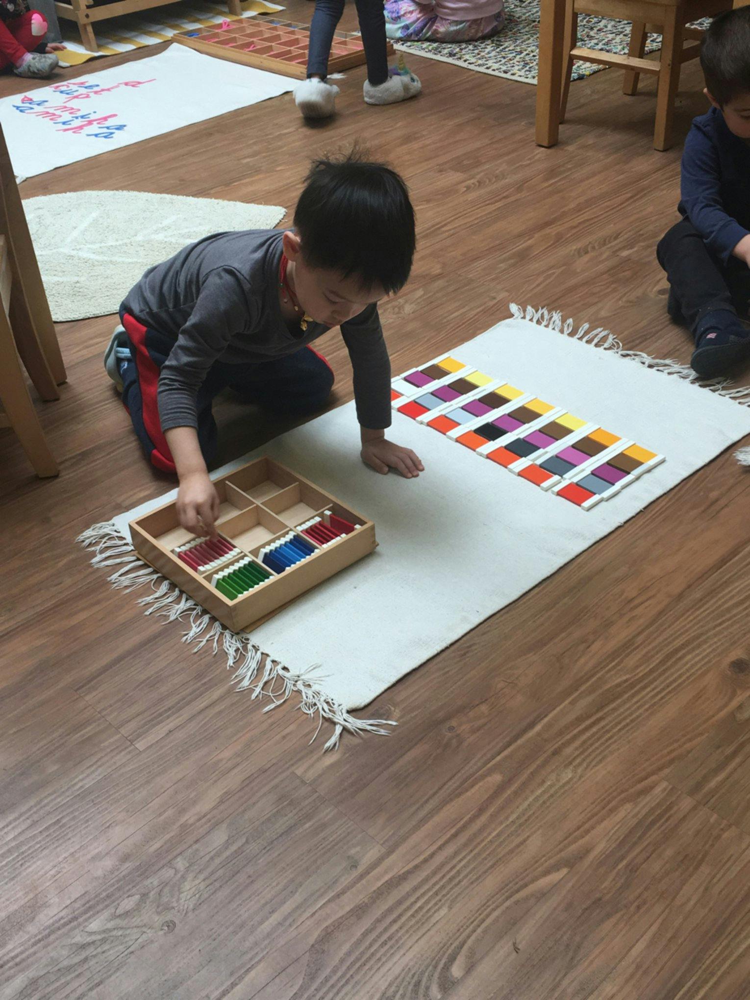 Child kneeling on floor, arranging colorful blocks from a wooden tray onto a mat