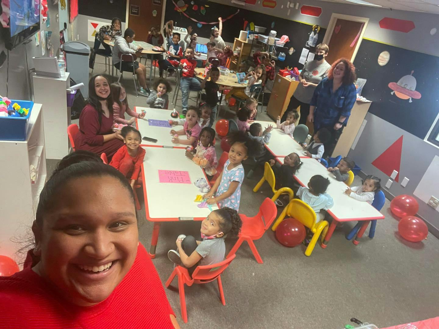 Person in red shirt smiling at camera with children and adults in a classroom