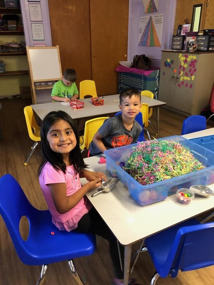 Children at tables in a classroom, one child smiling at the camera