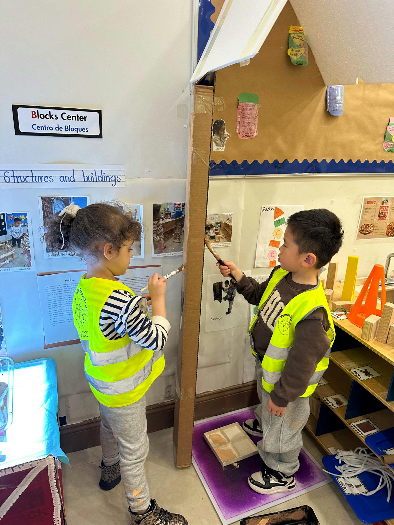 Two children wearing neon safety vests painting a large cardboard structure