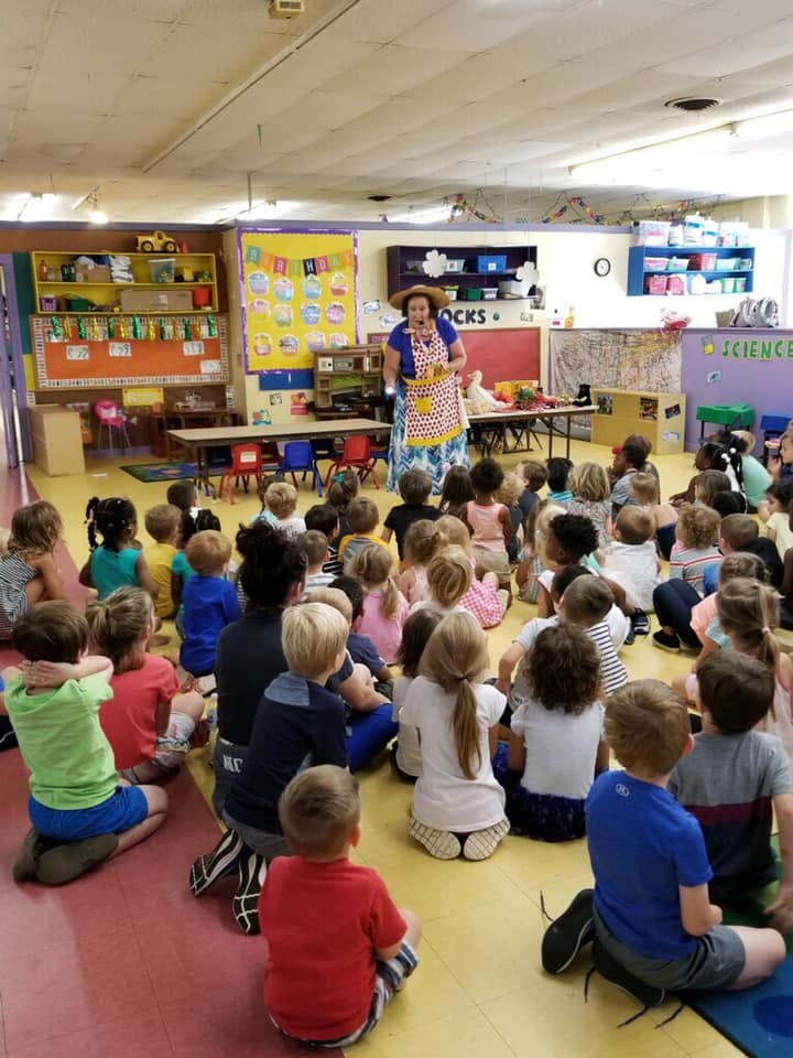 Person in an apron and hat speaking to a group of children in a classroom