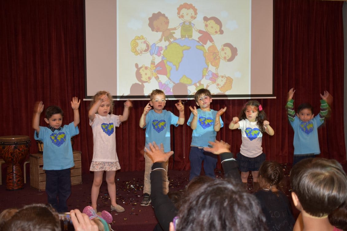 Children on a stage with hands raised, a projection of children around a globe behind them