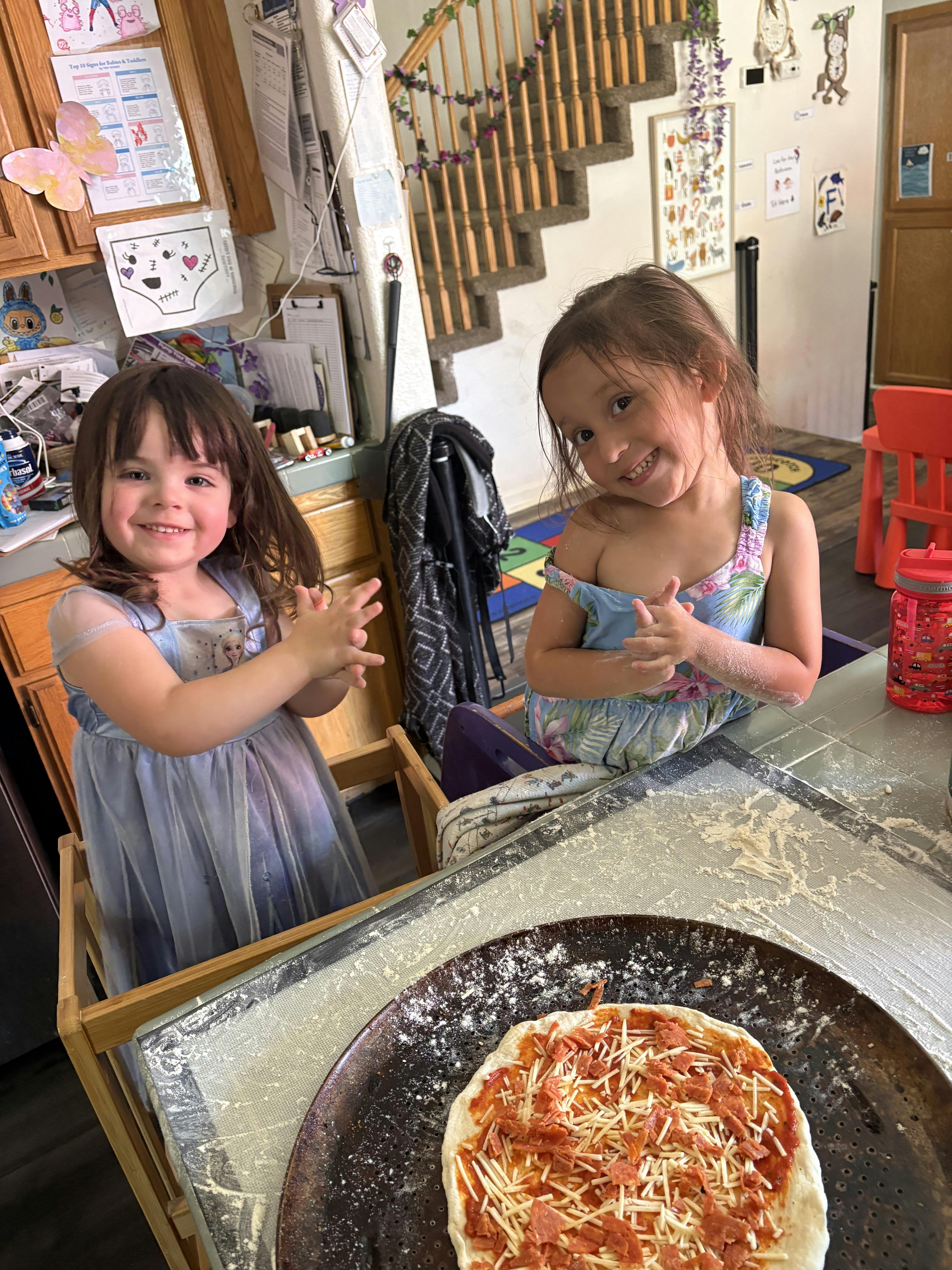 Two children at a table preparing a pizza with flour on their hands