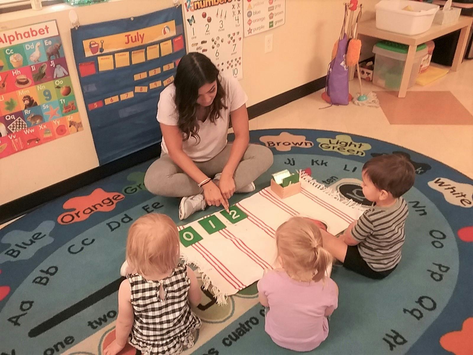 Person sitting on floor with three children, pointing at numbers on a mat