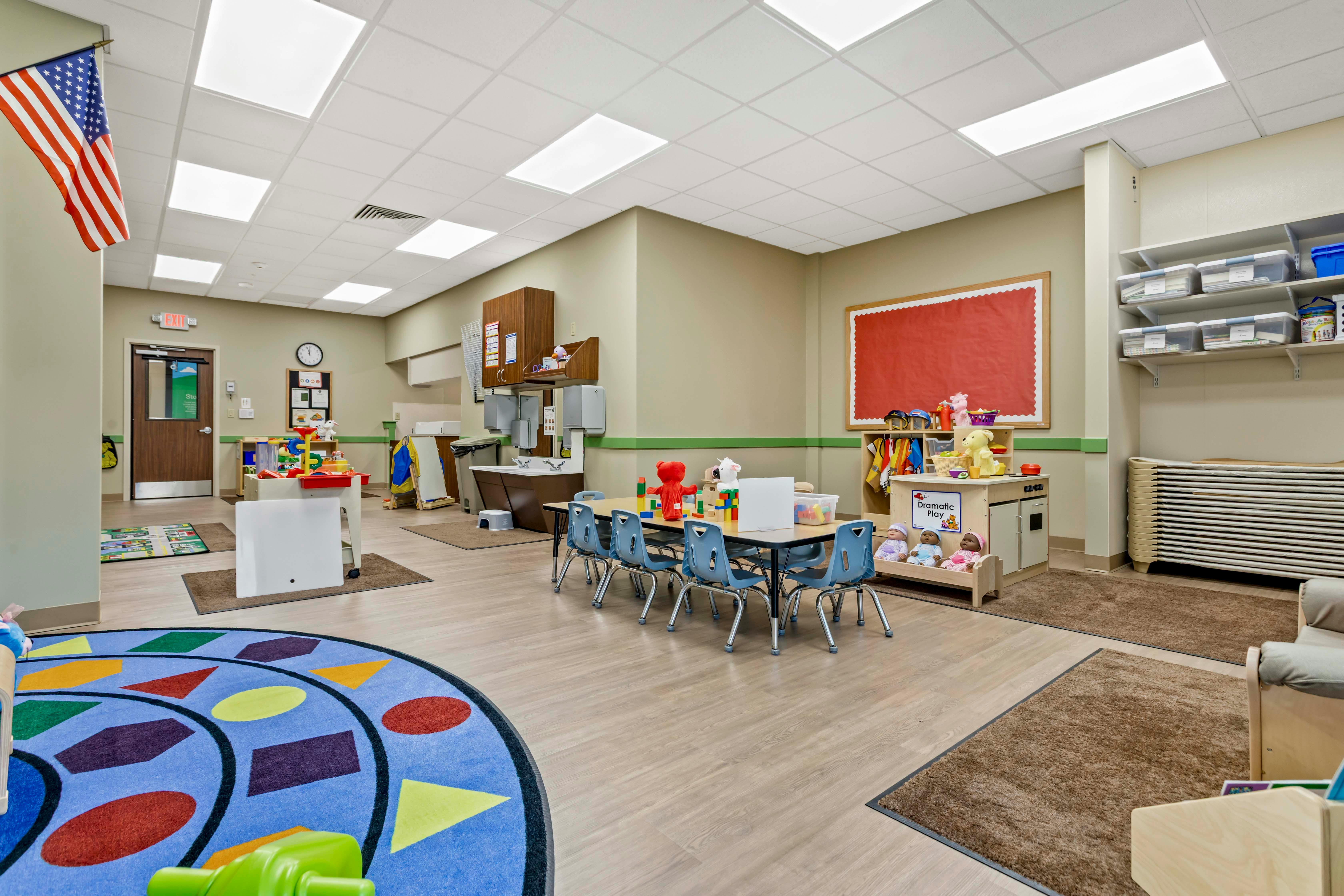 An empty childcare classroom with small tables and chairs, toys, and a flag