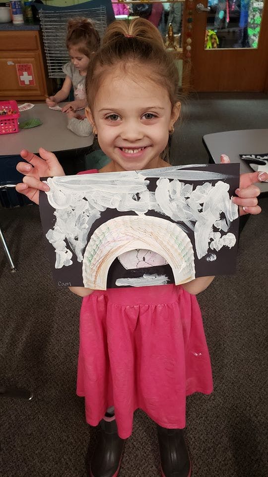 Child holding up a paper craft with a rainbow and white clouds