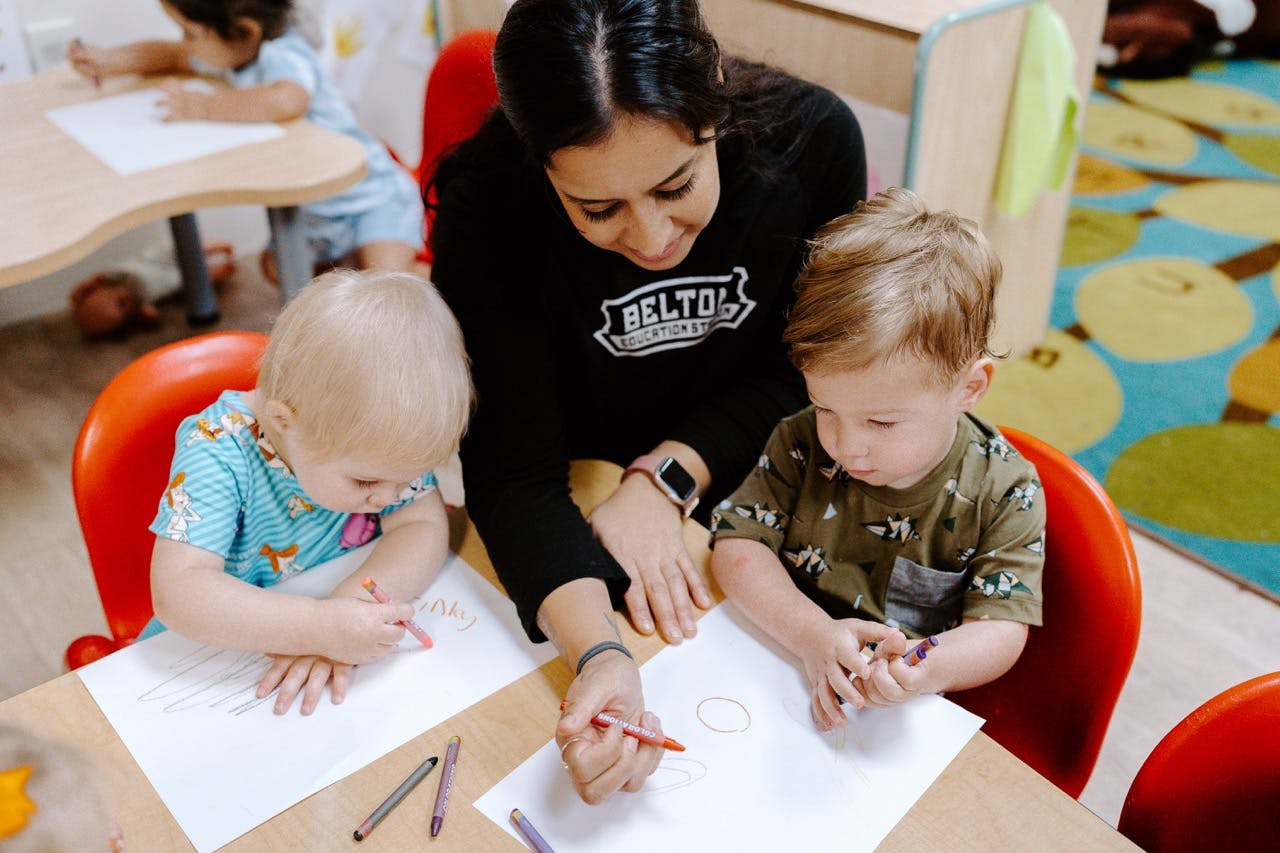 Person helping two children draw with crayons in a classroom