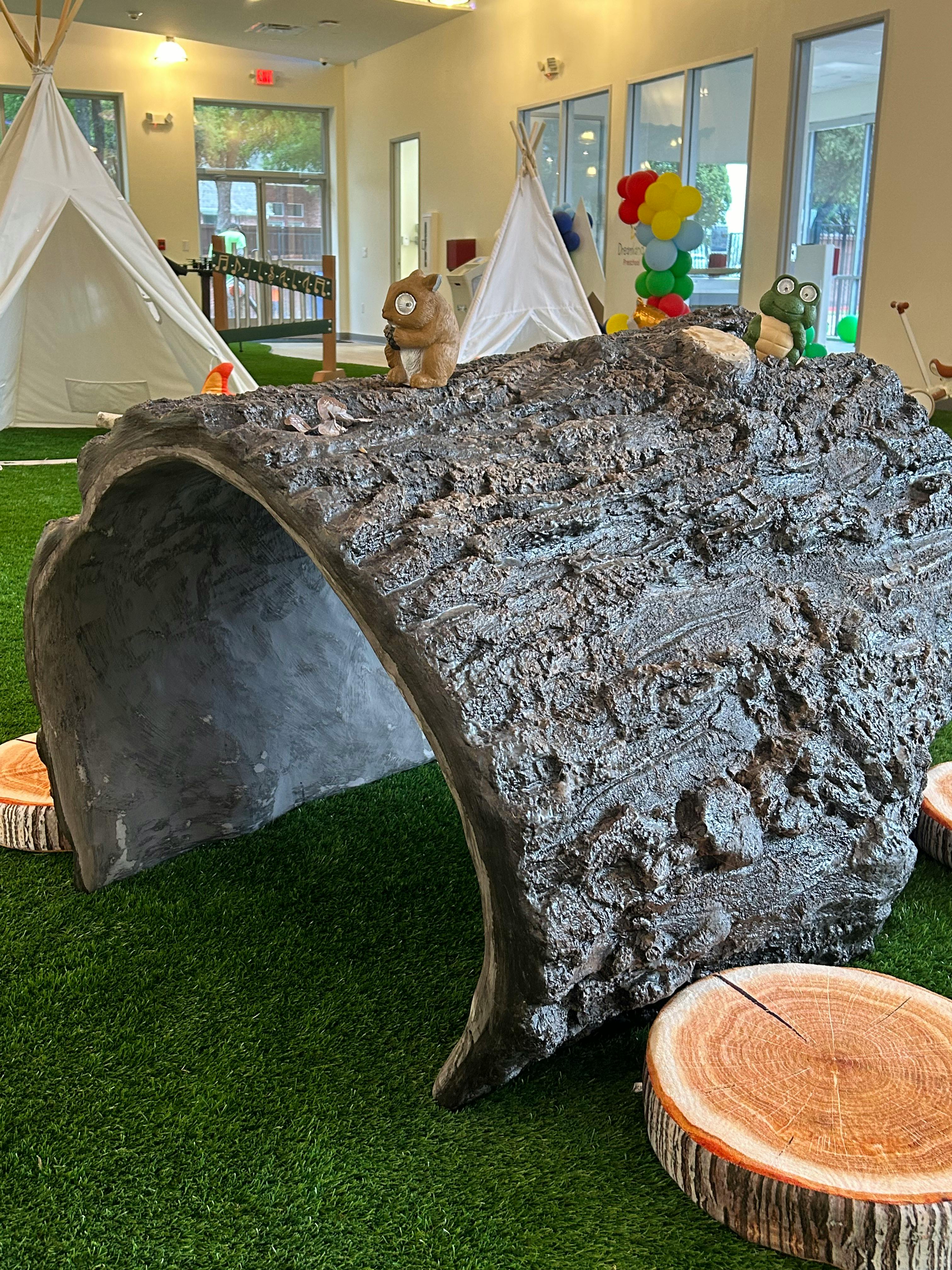 Large faux log tunnel on green turf with teepees and stuffed animals in background
