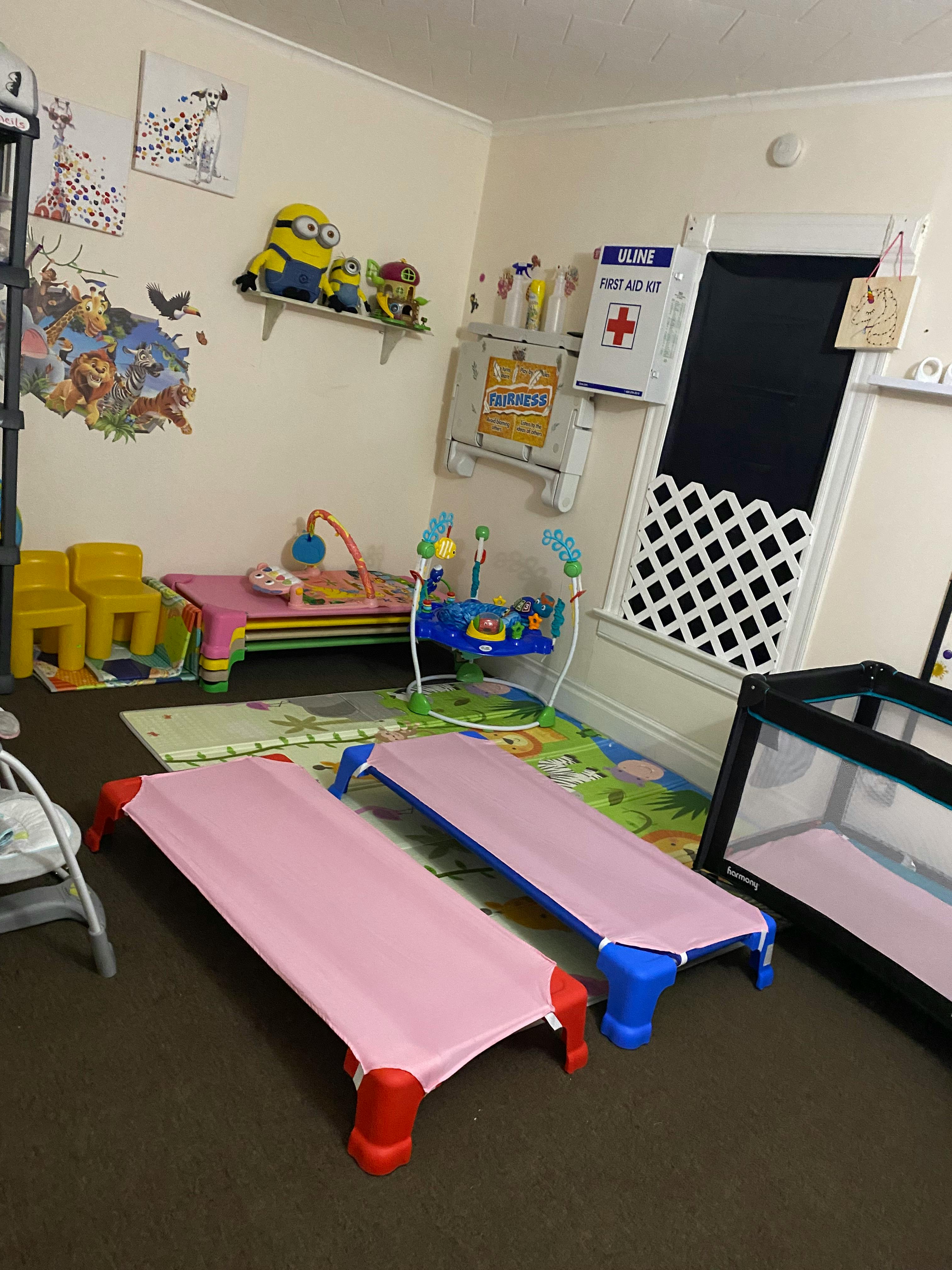 Childcare room with two pink cots, play mat, toys, and a first aid kit on the wall