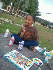 Child sitting on ground with paint, brushes, and a colorful canvas