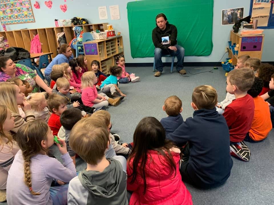 Adult sitting on a chair showing a book to a group of children in a classroom