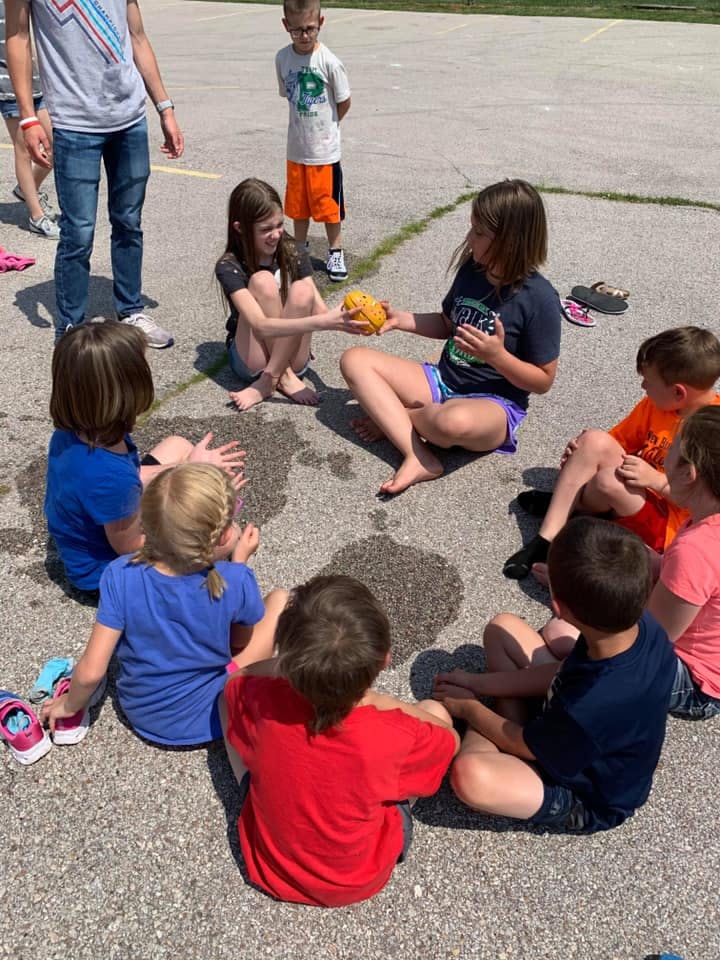Children sitting in a circle on pavement, one child passing an object to another