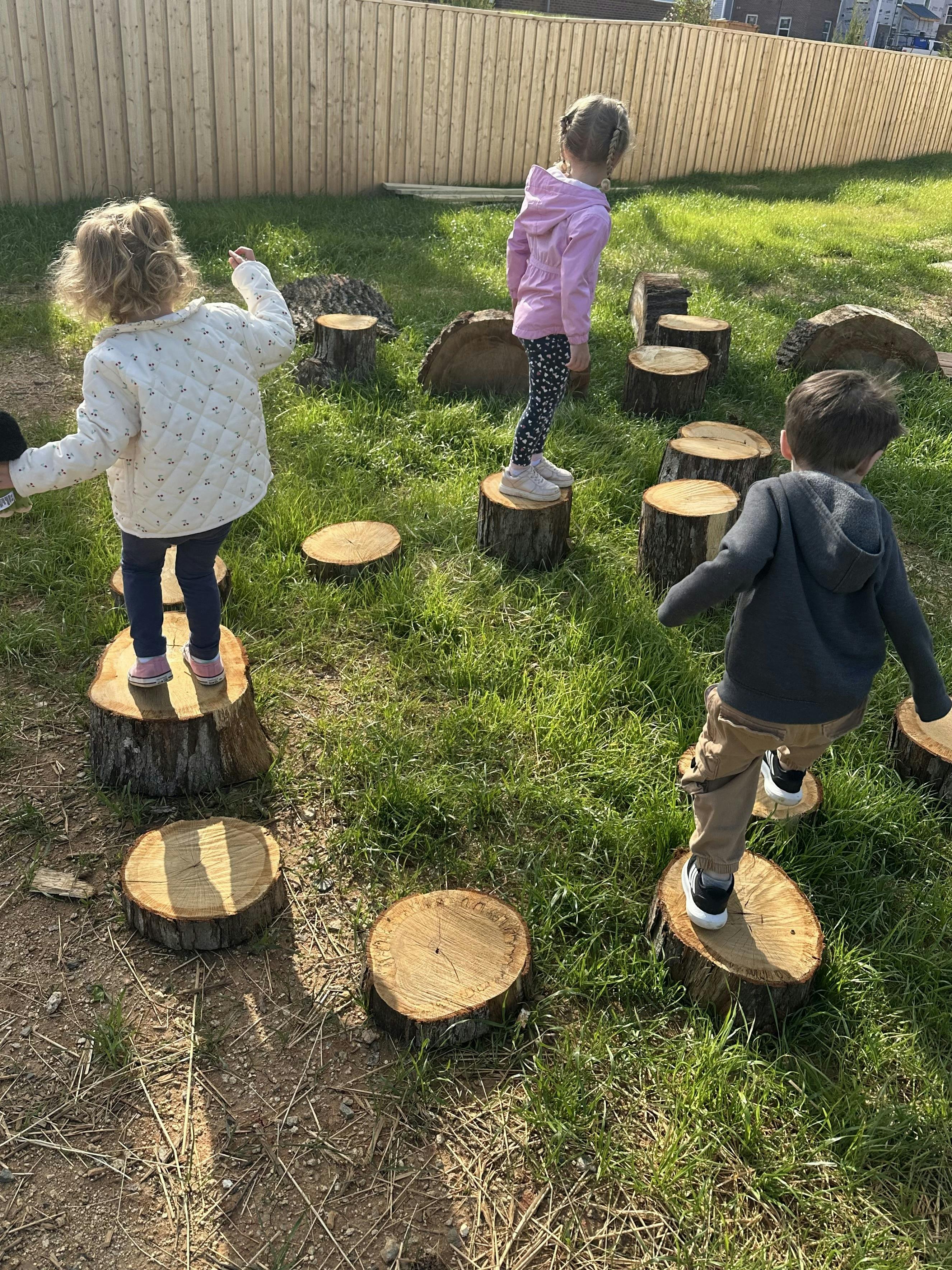 Three children balancing on tree stumps in a grassy area
