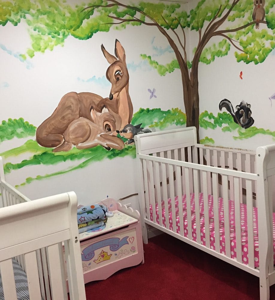 Two white cribs in a room with a wall mural of deer and a tree