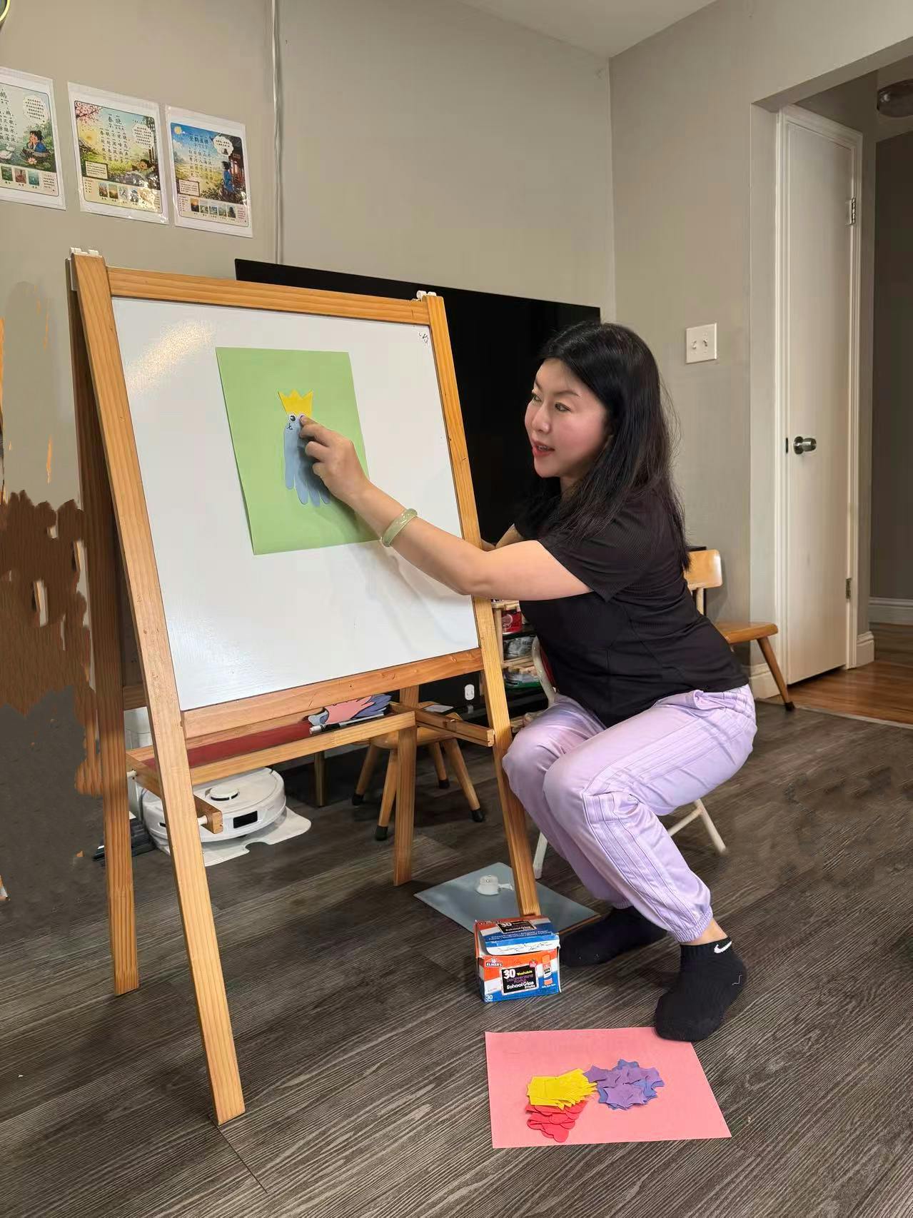 Person squatting and gluing a paper crown onto a blue paper cutout on an easel