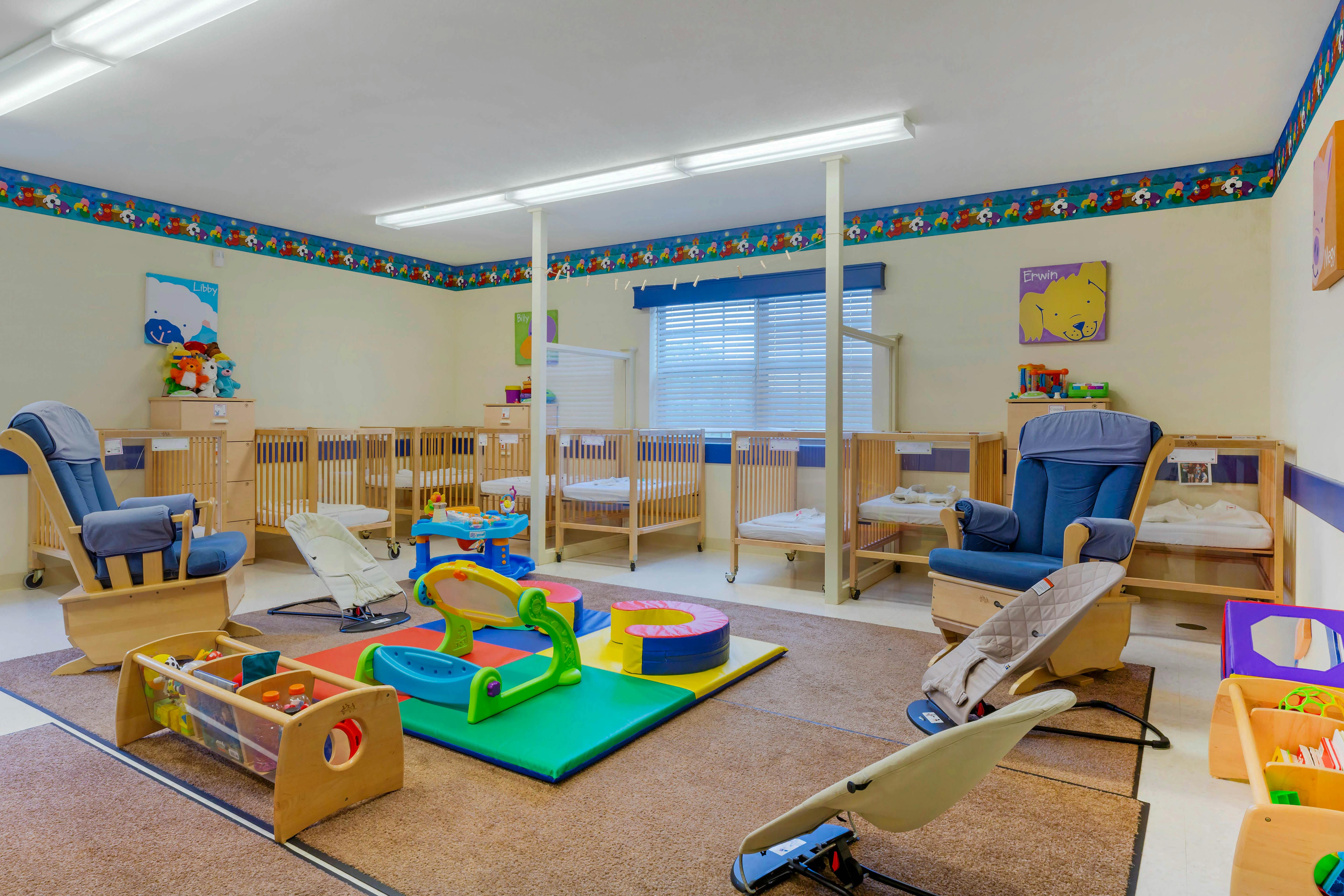 A brightly lit nursery with multiple cribs, rockers, playmats, and toys