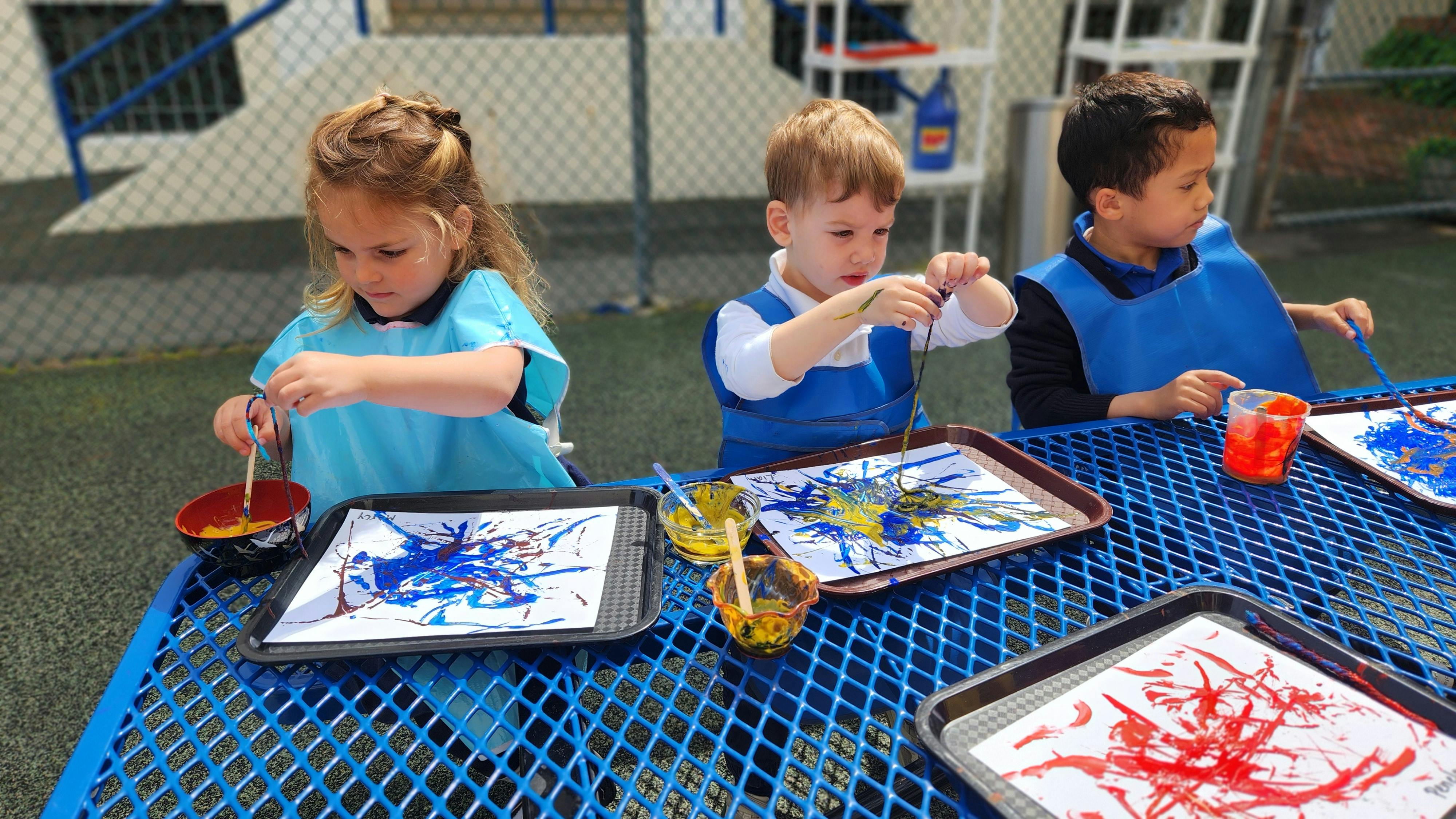 Three children painting at a blue outdoor table