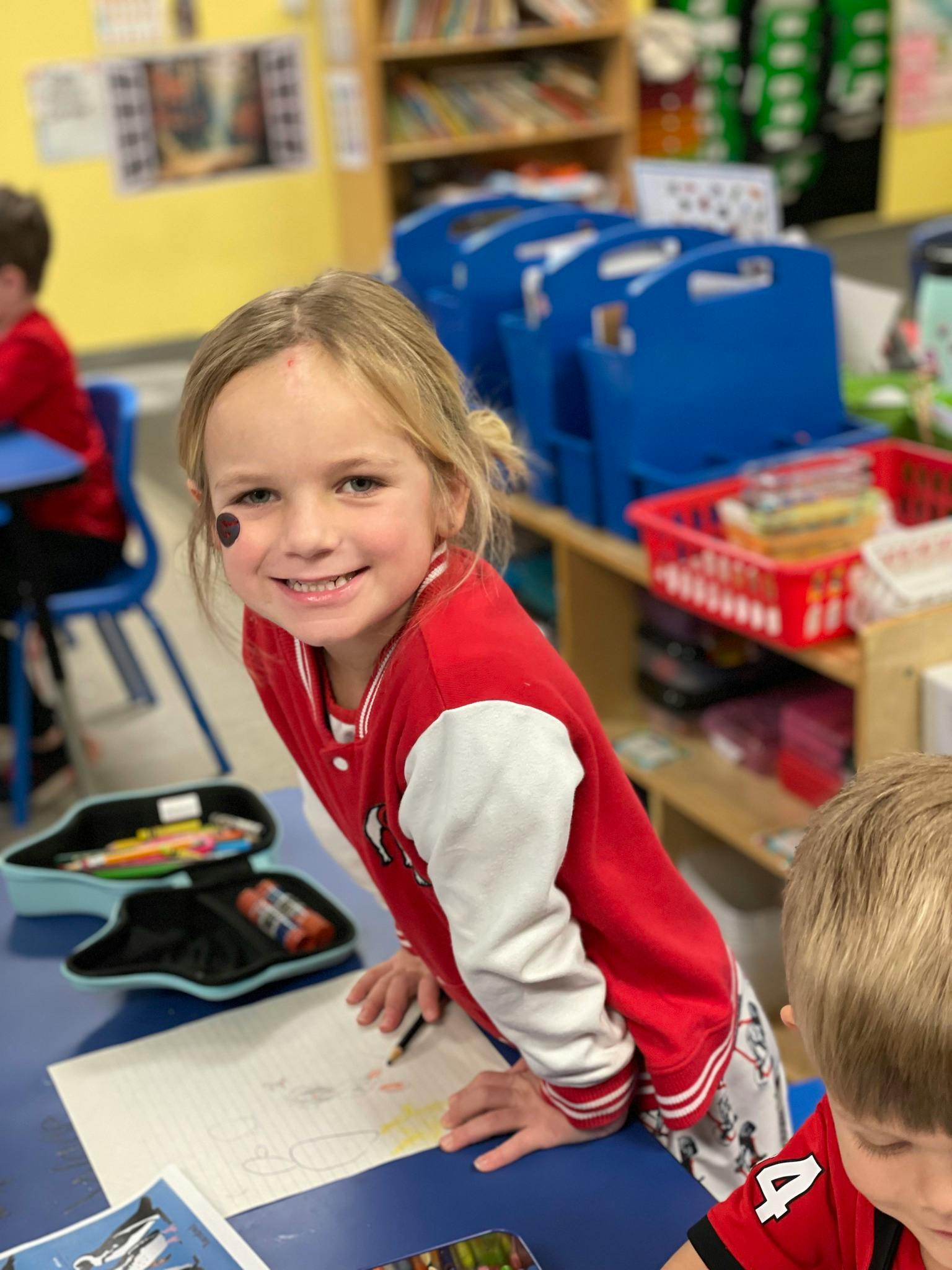Child in a classroom smiling at the camera while drawing
