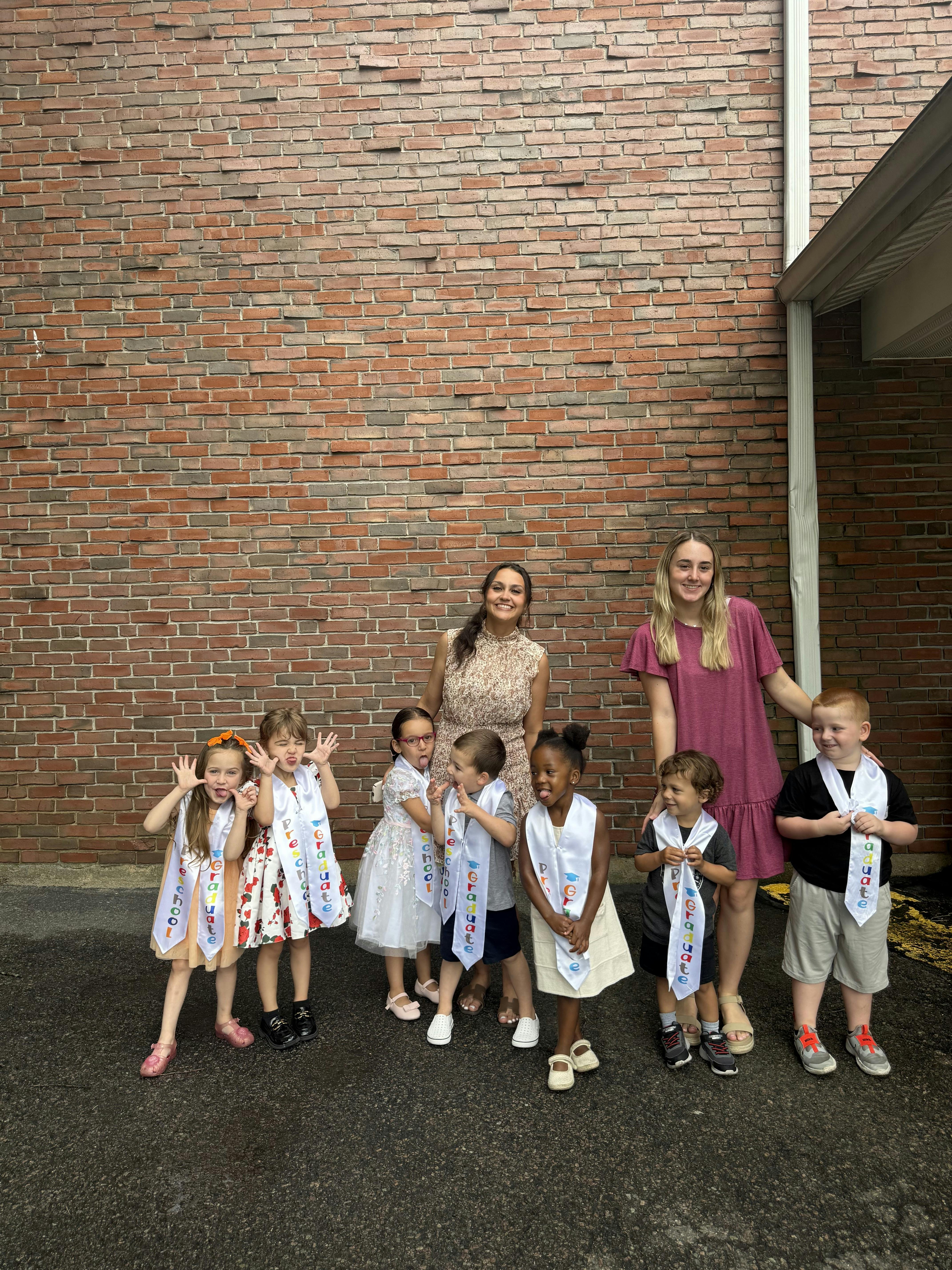 Group of children wearing preschool graduate sashes with two adults