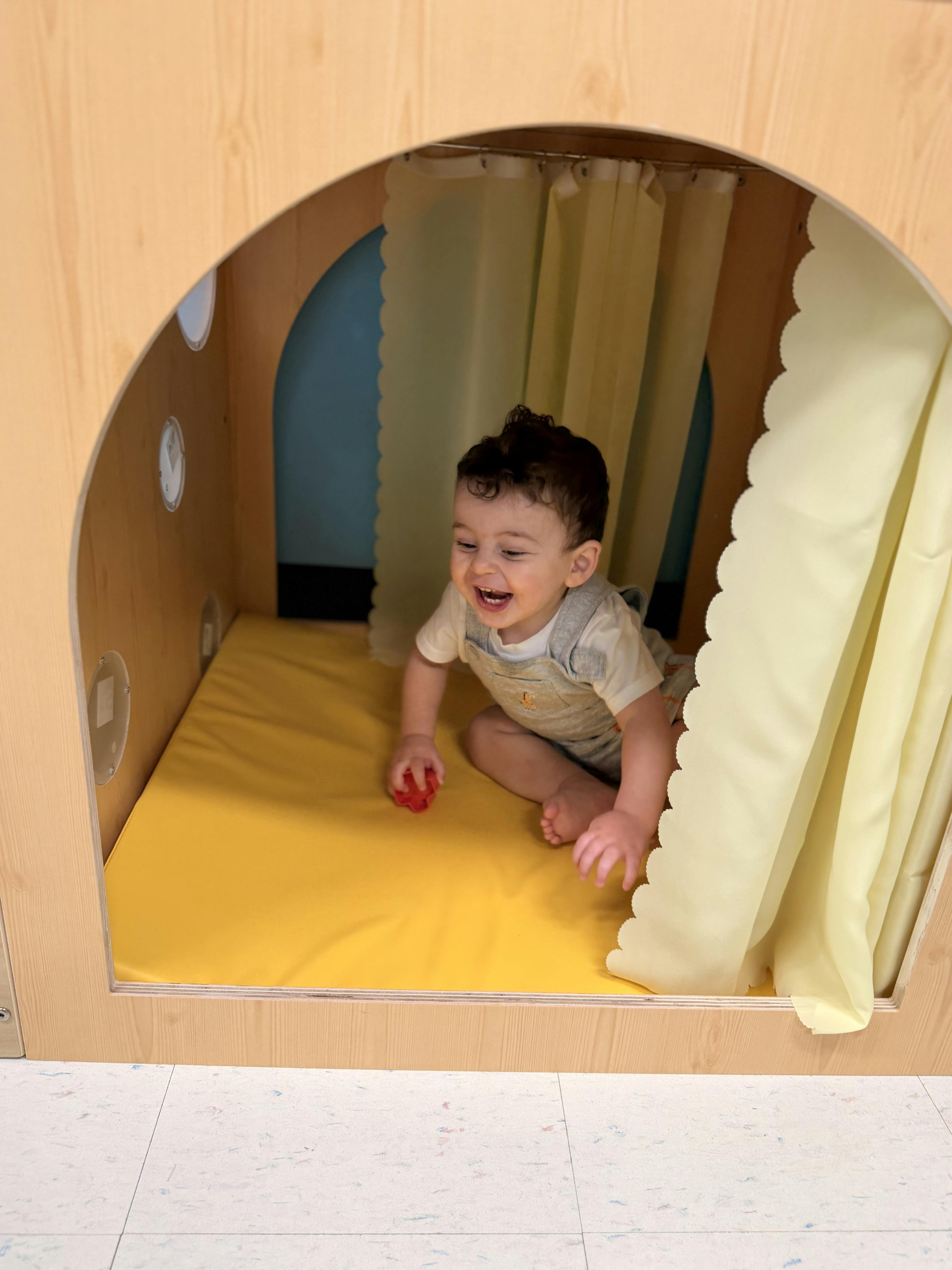 Child sitting inside a wooden play structure with yellow curtains