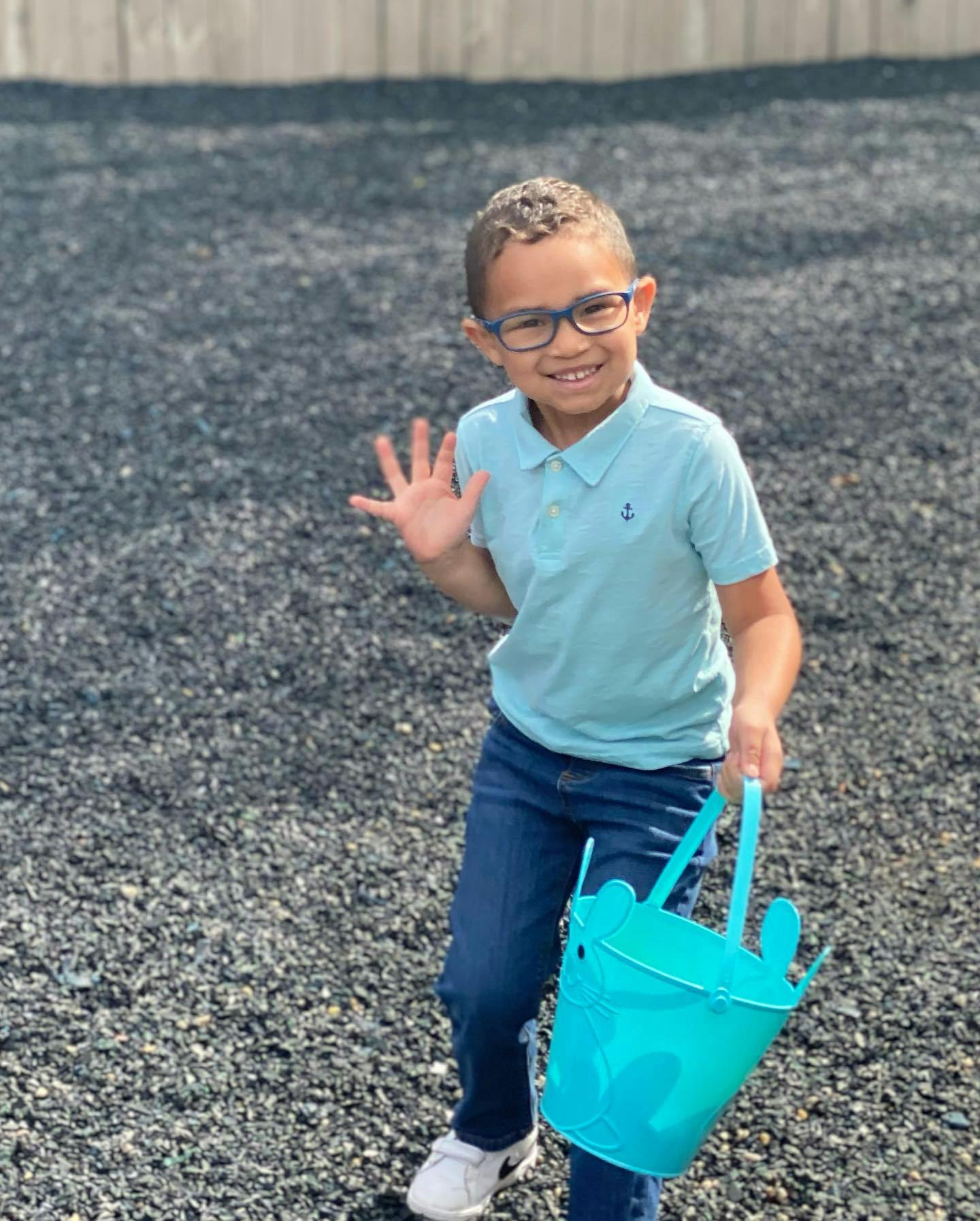 Child wearing glasses, holding a blue bucket, and waving
