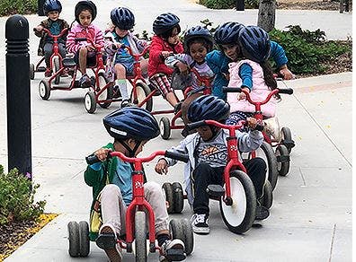Children wearing helmets riding tricycles in a line
