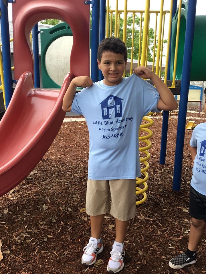 Child pulling up a t-shirt with "Little Blue Academy" text on a playground