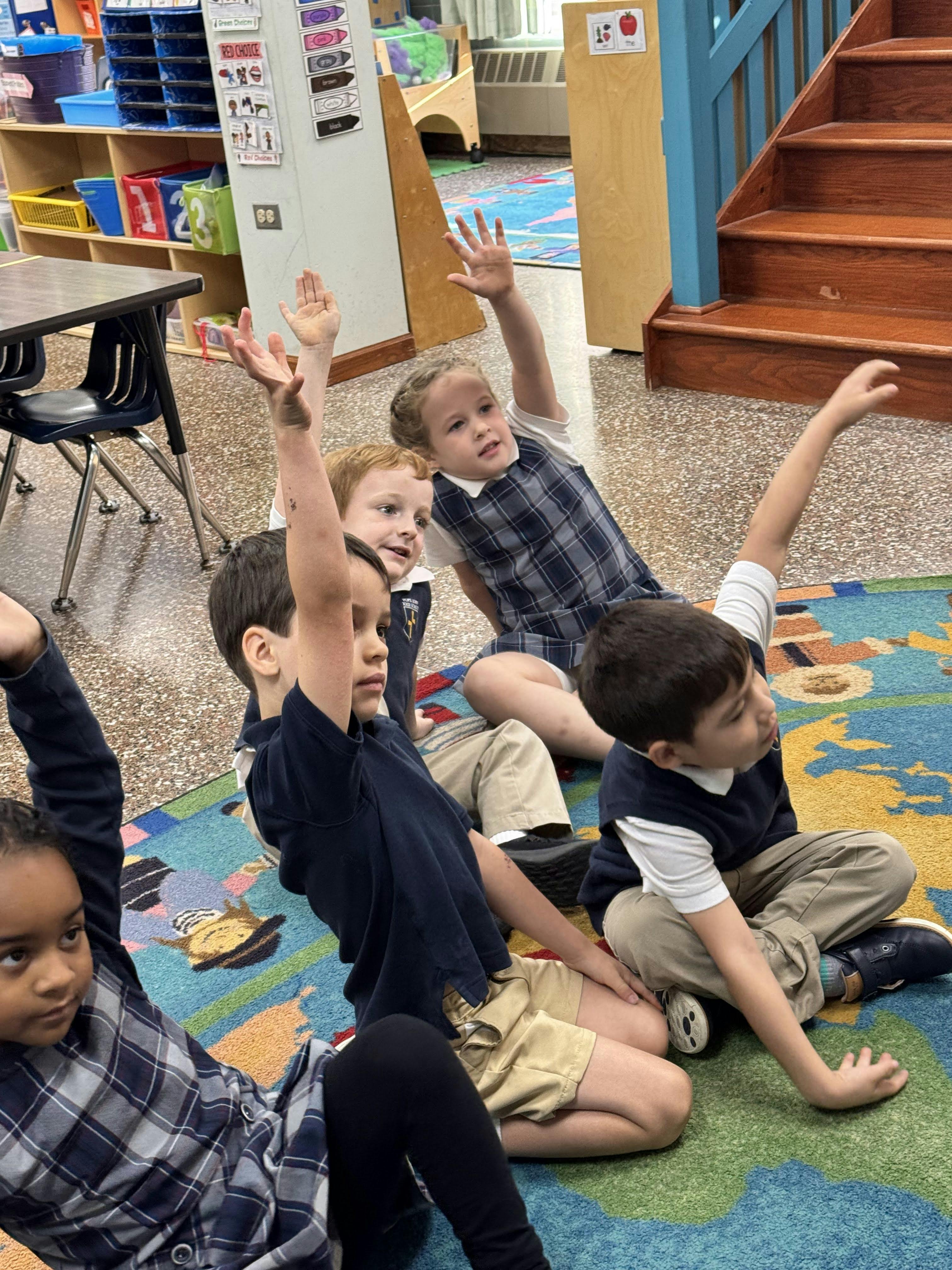 Children sitting on a rug with some raising their hands in a classroom