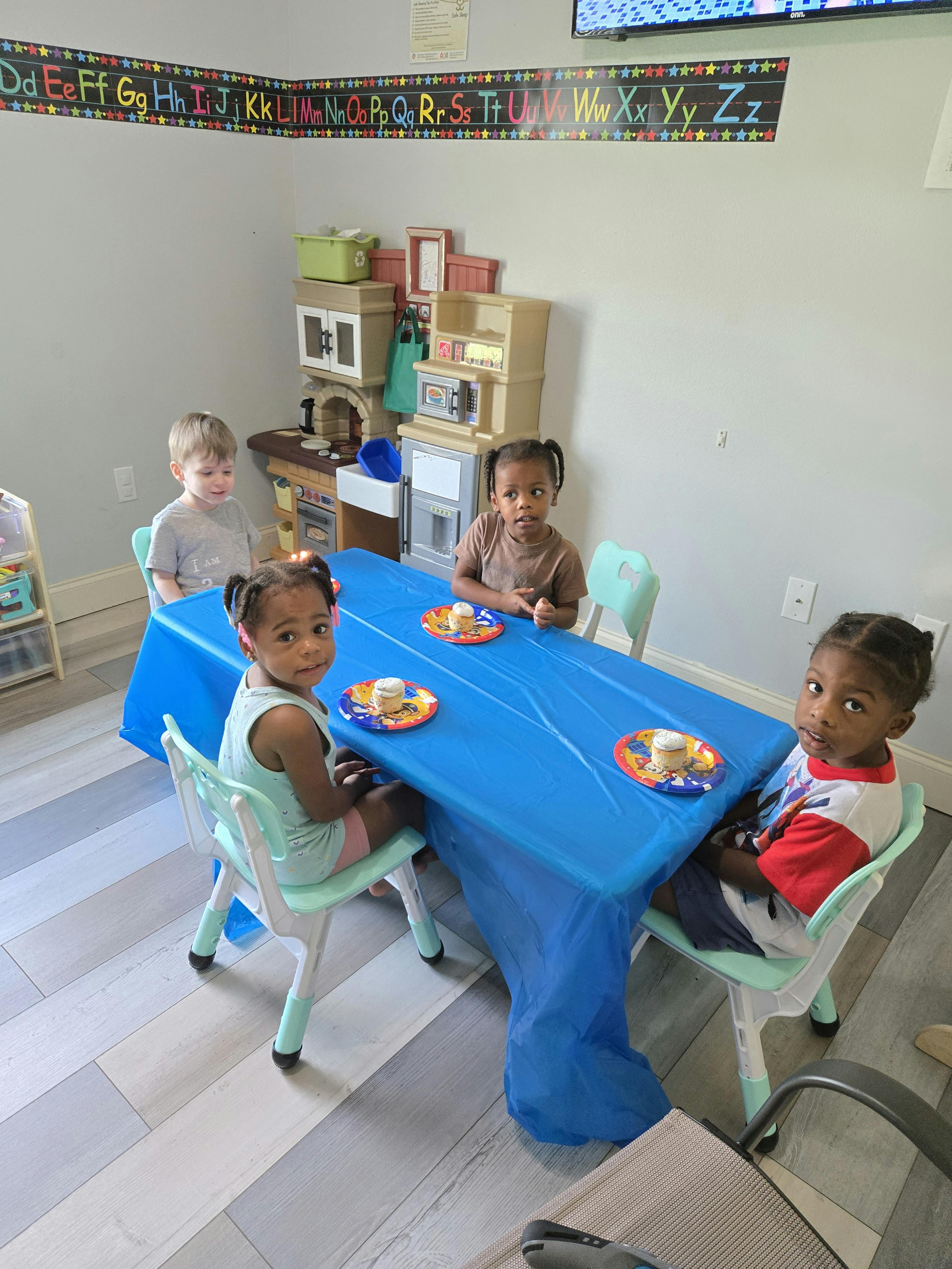 Four children sitting at a blue table with plates in a classroom