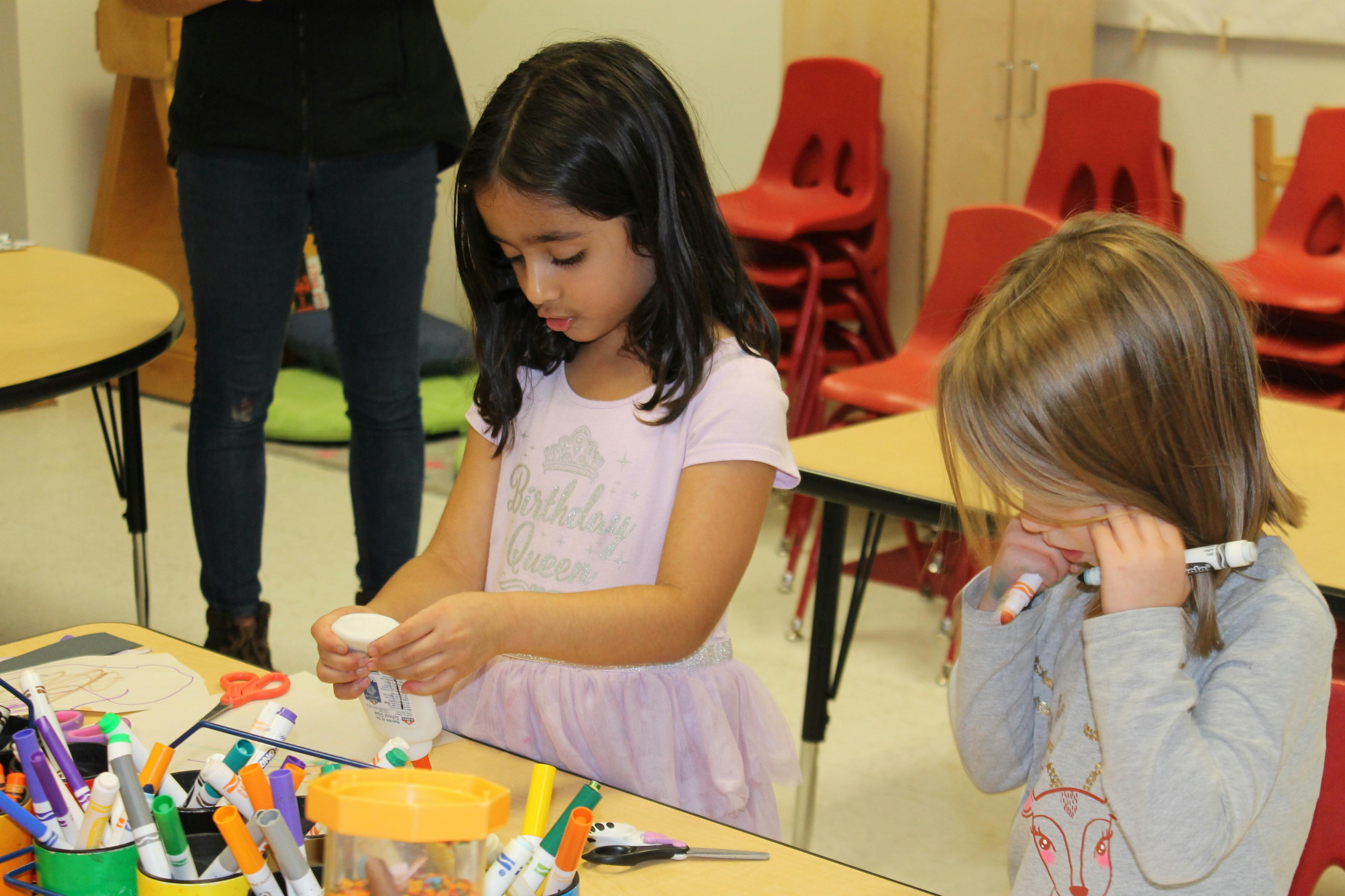 Two children at a table, one squeezing a bottle, the other holding a marker