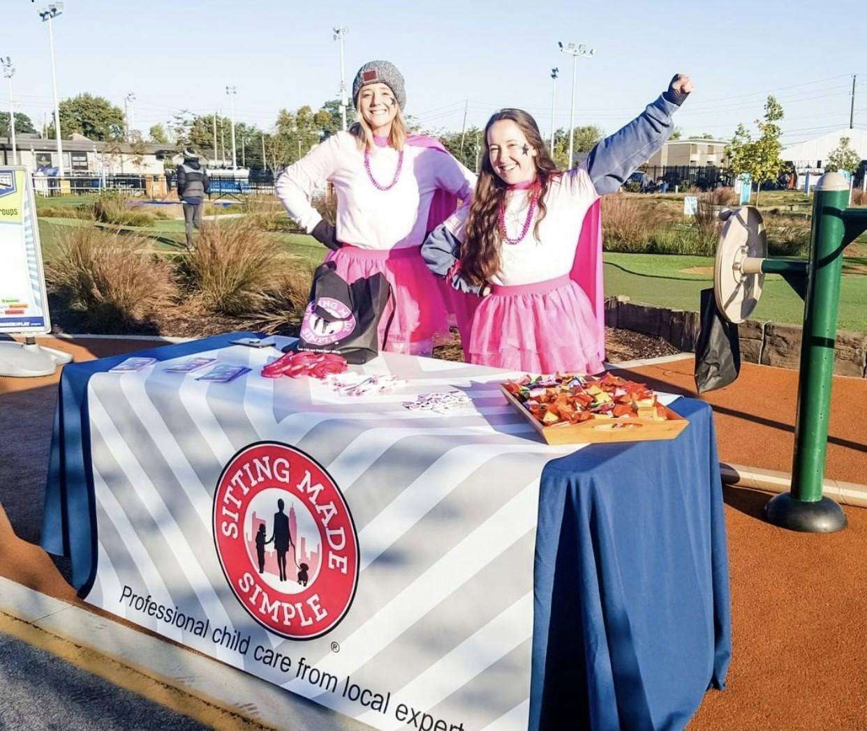 Two people in pink outfits standing behind a table with a "Sitting Made Simple" sign