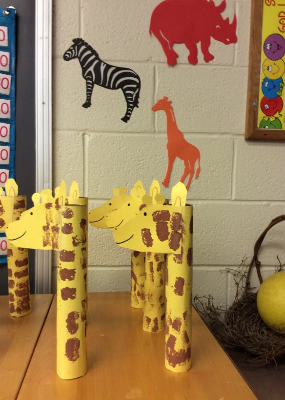 Giraffe crafts made from paper tubes on a table, with animal cutouts on the wall