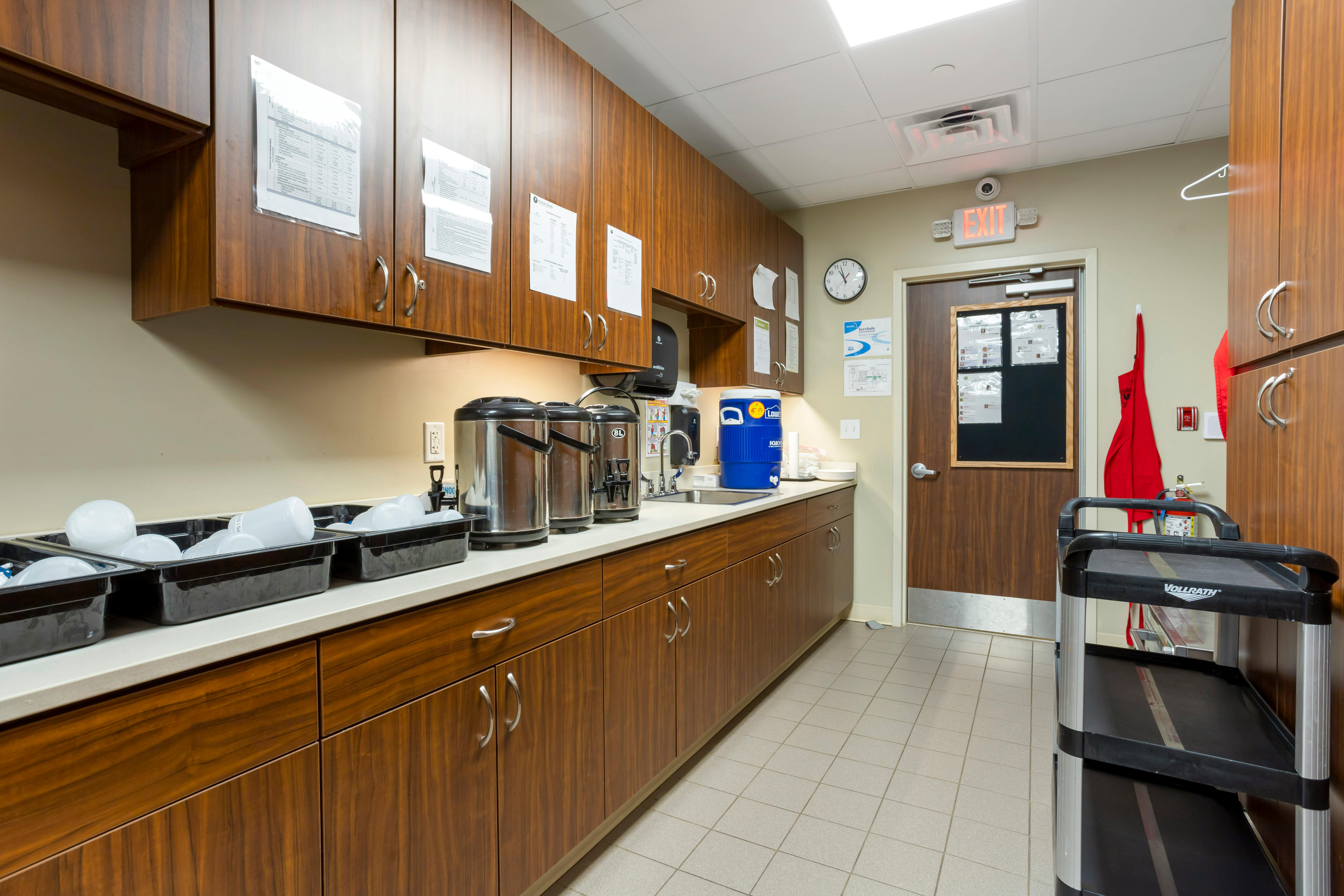 Hospital kitchen with wooden cabinets, metal carafes, a sink, and a red apron