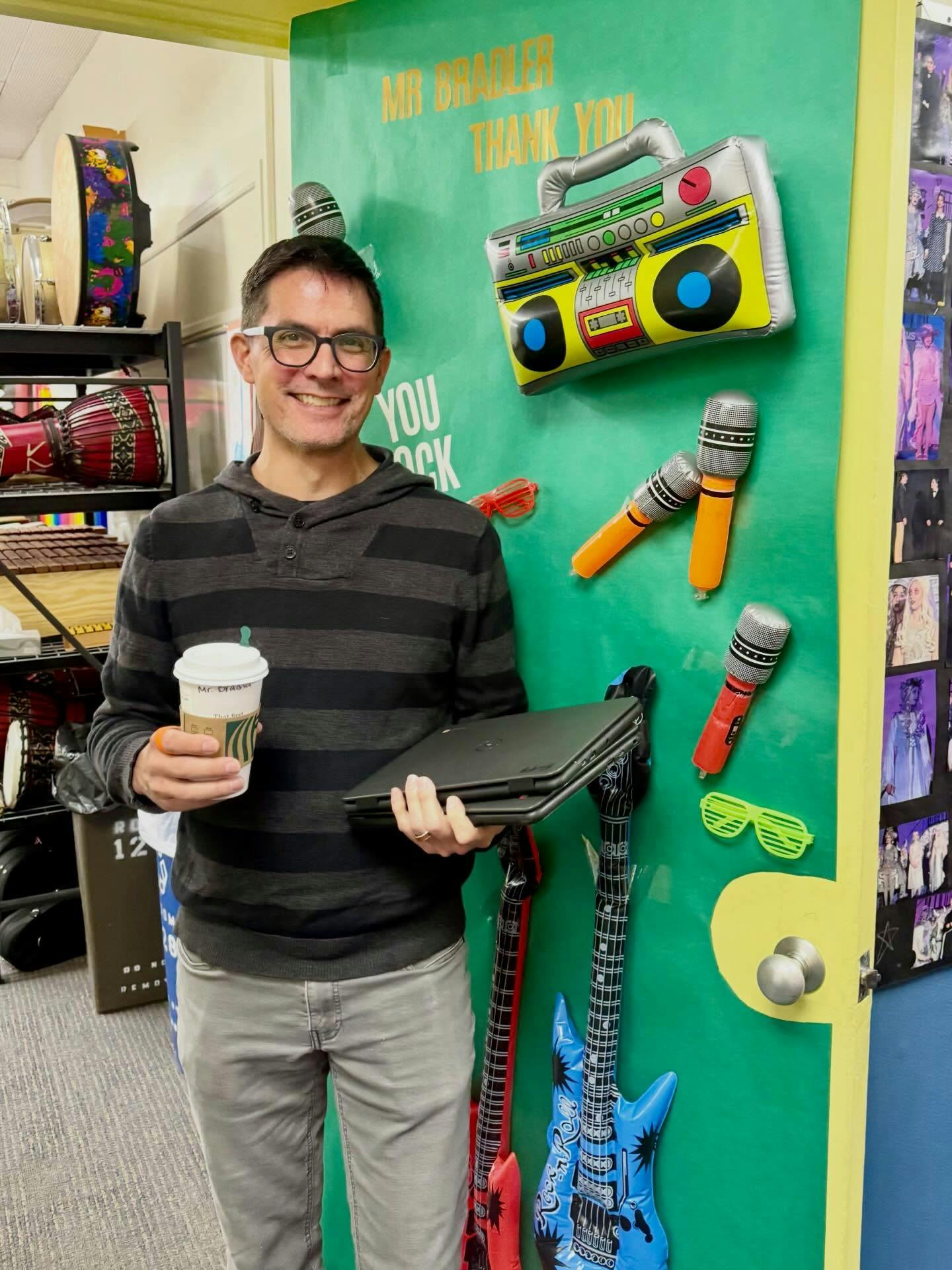 Smiling person in glasses holding a coffee and laptop next to a door with inflatable musical decorations