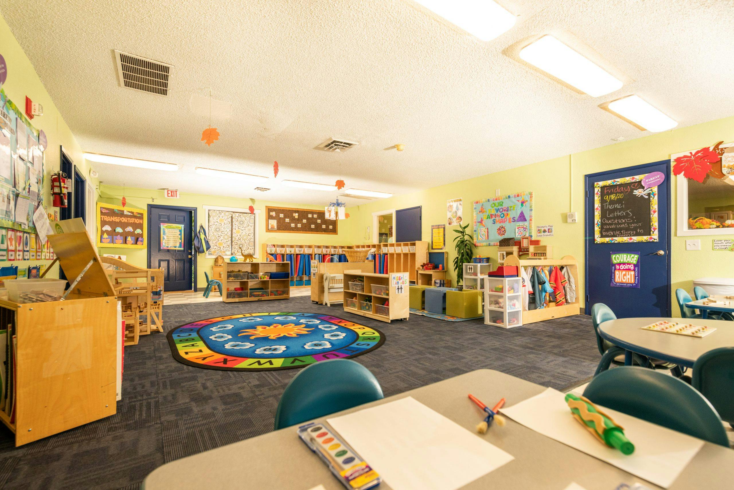 A colorful preschool classroom with tables, chairs, shelves, and a circular alphabet rug