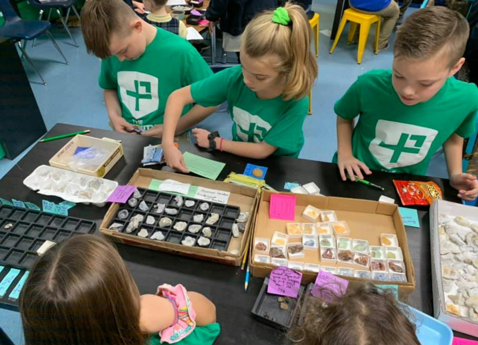 Children in green shirts examining rocks and minerals on a table