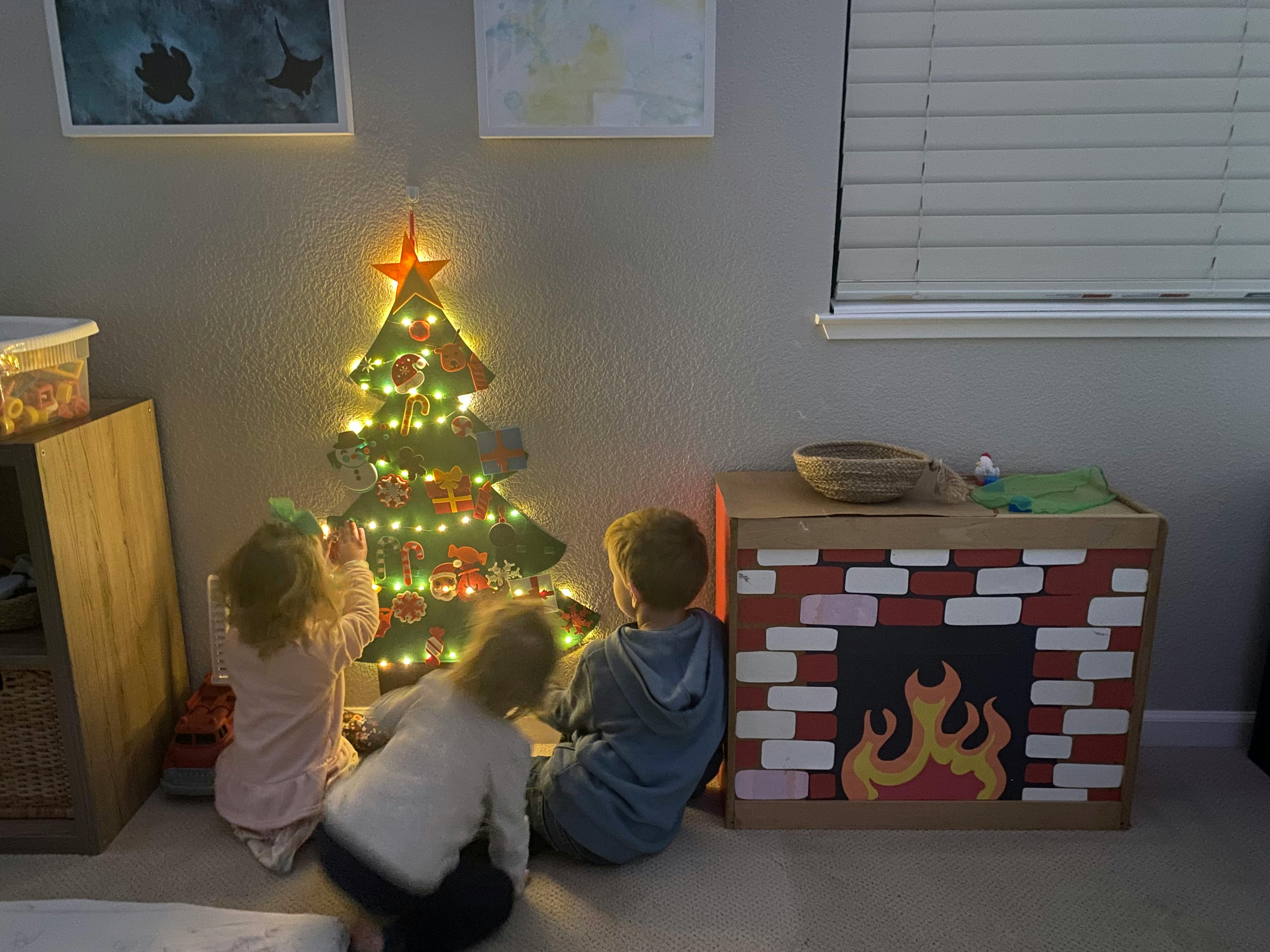 Three children sitting on the floor in front of a light-up felt Christmas tree