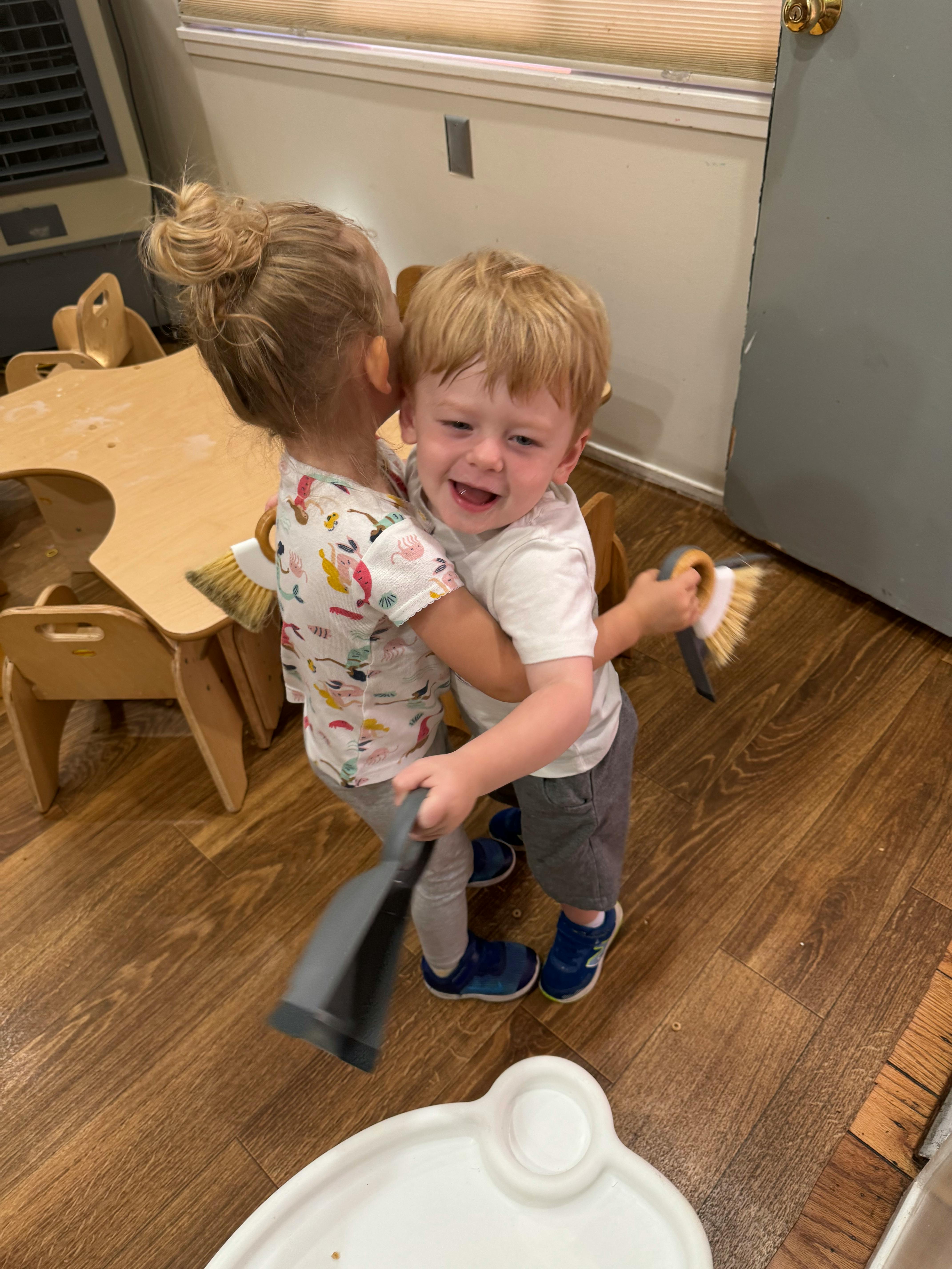Two children hugging, one holding a brush and the other a dustpan
