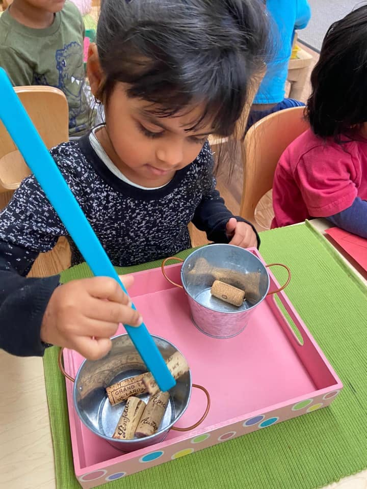 Child using a blue tool to move corks between two small metal buckets on a tray
