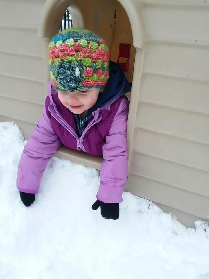 Child in a winter coat and hat looking out of a playhouse window into snow