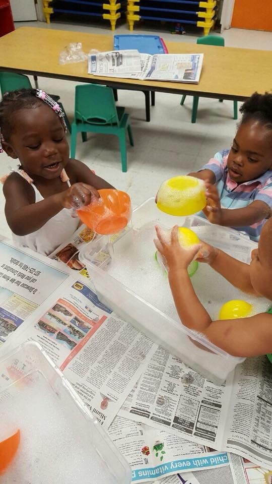 Three children playing with soapy water and colorful bowls in a classroom