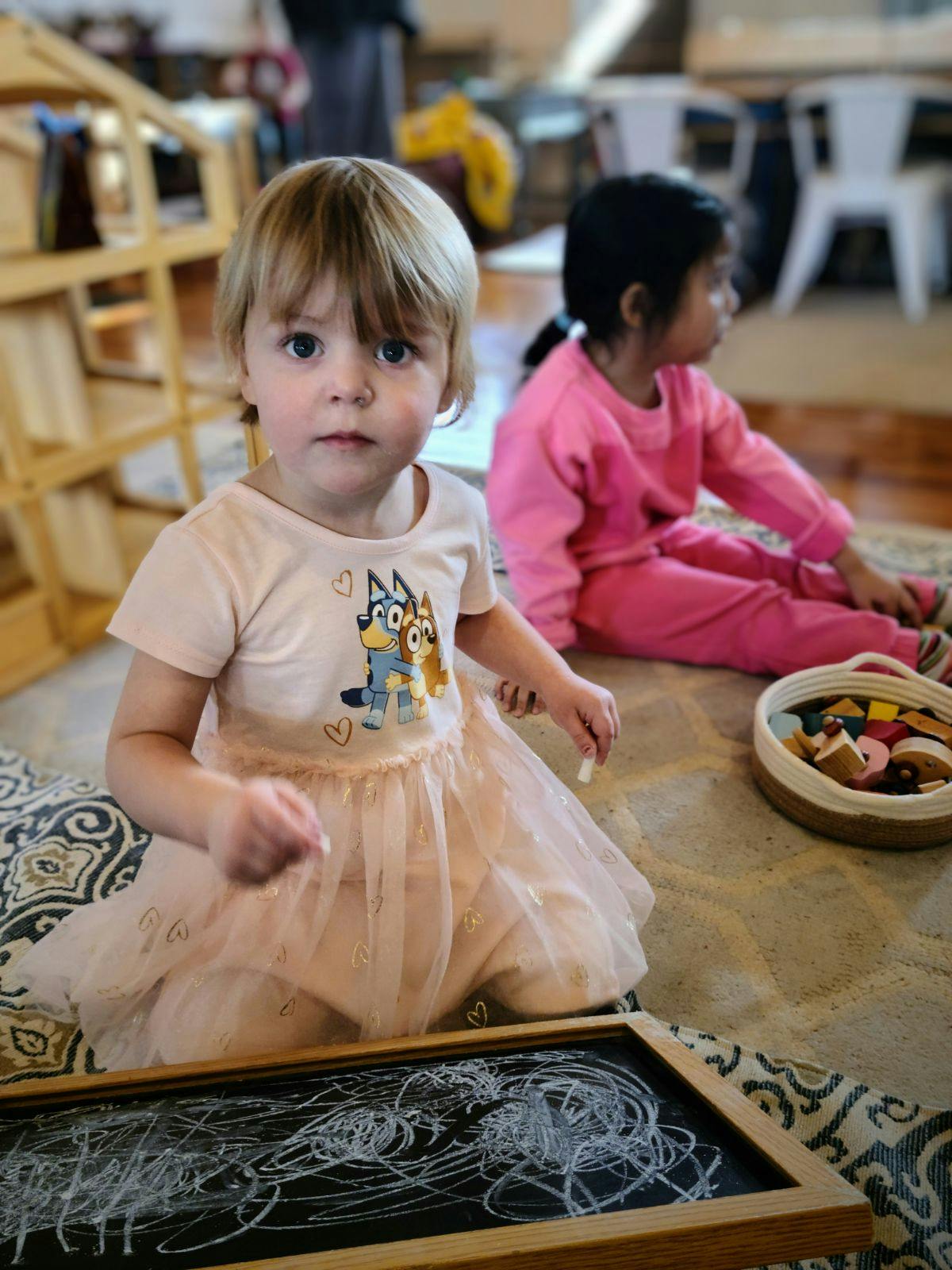 Child in a dress kneeling on a rug with a chalkboard in front