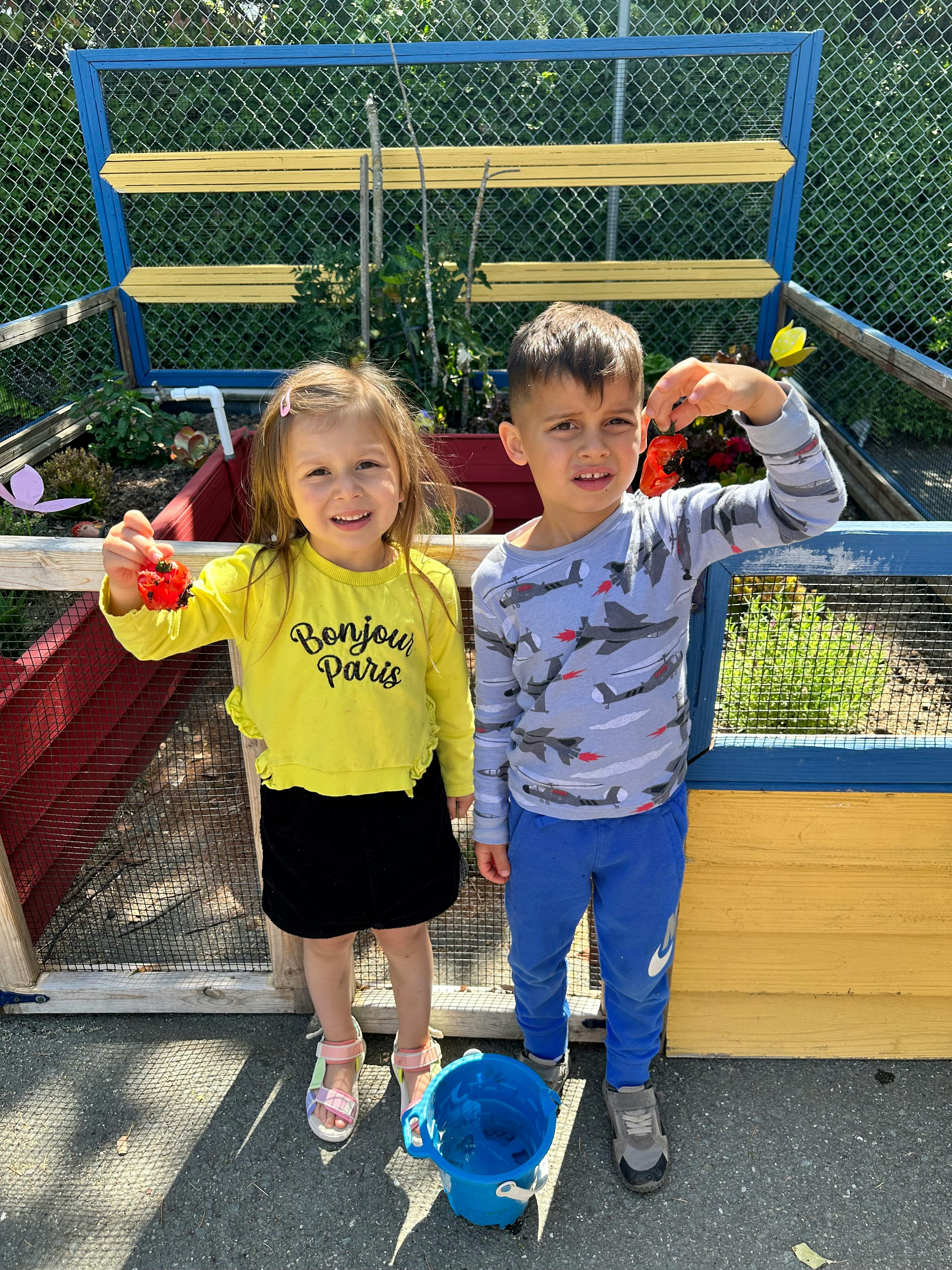 Two children standing outdoors, one holding a red object