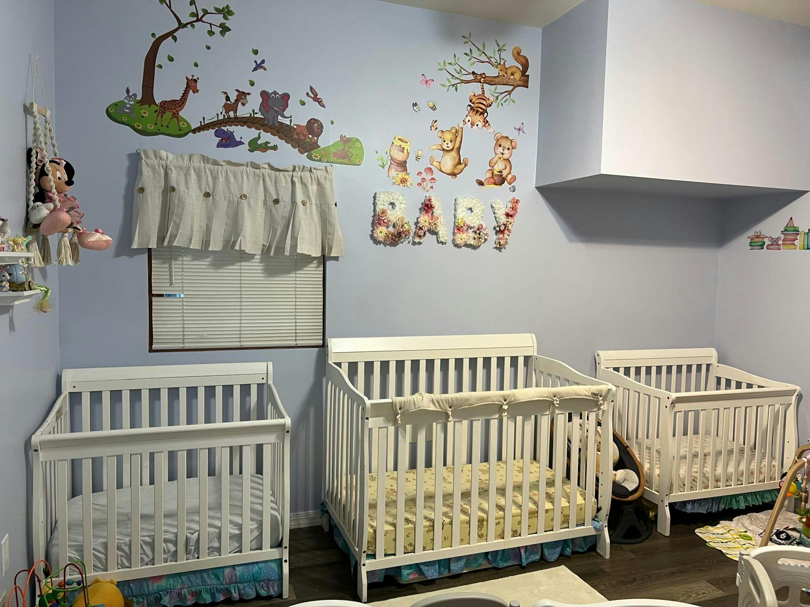 Three white cribs in a room with wall decals of animals and the word "BABY"