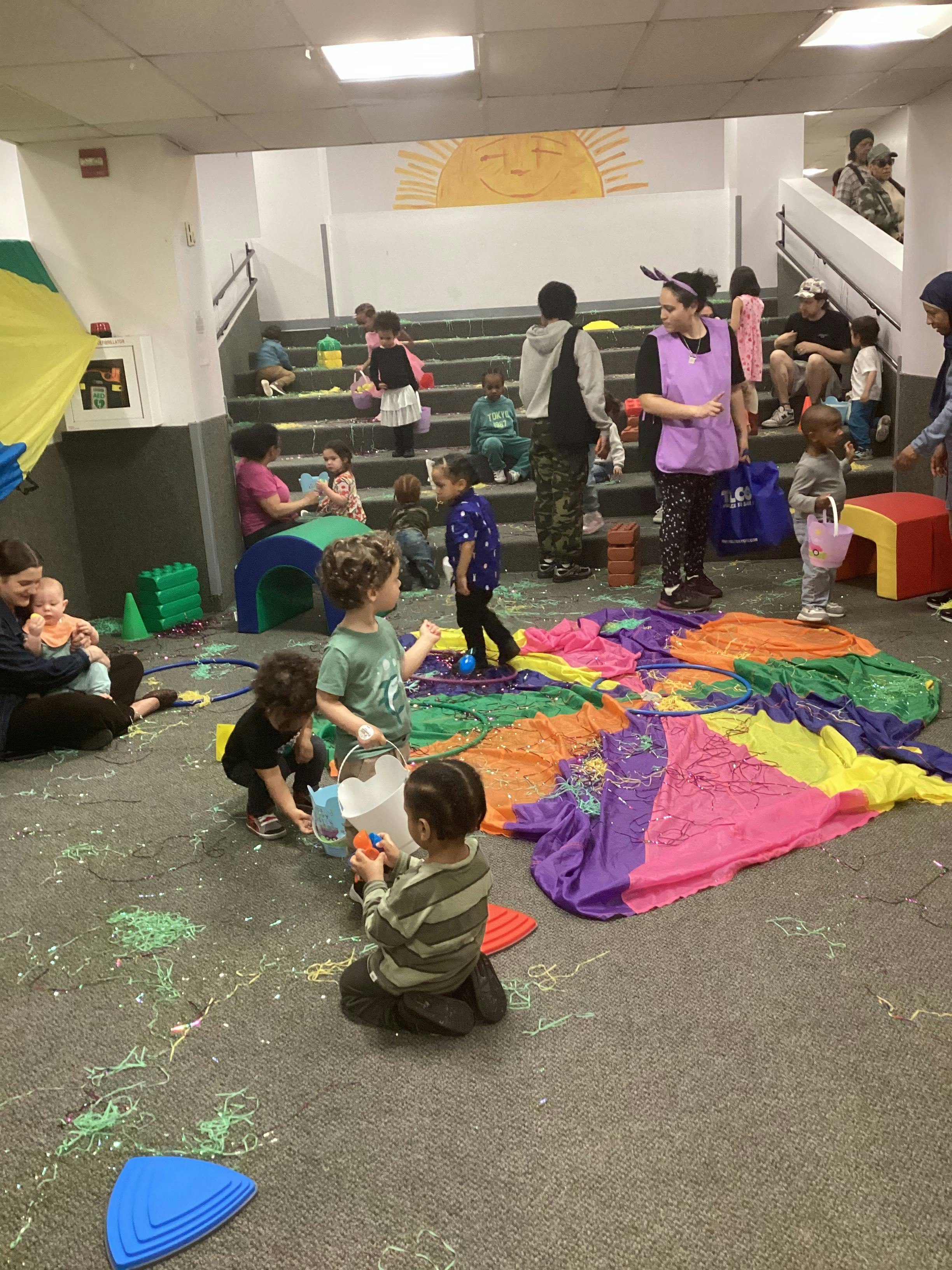 Children and adults gather around a colorful parachute and confetti on the floor