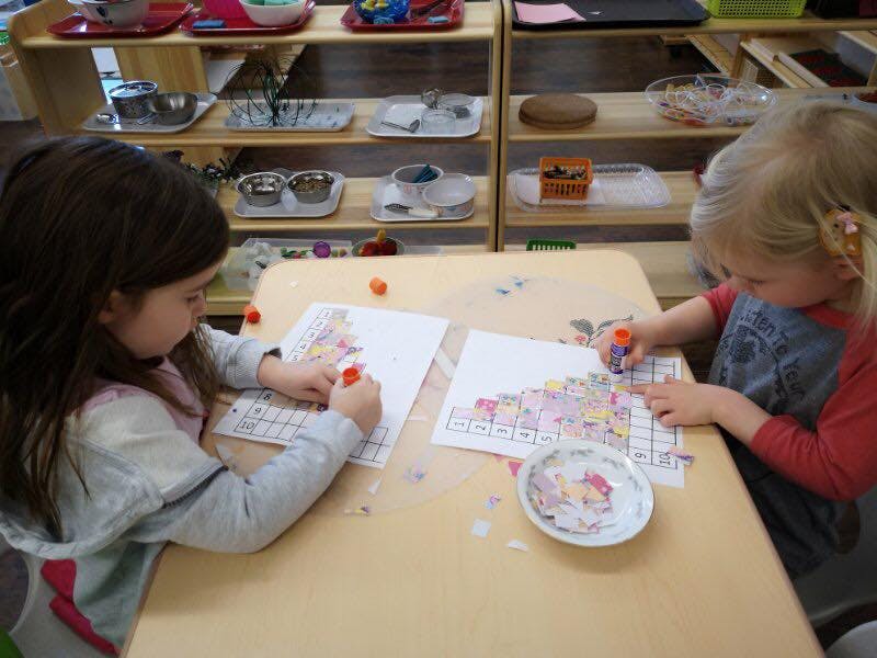 Two children gluing paper cutouts onto sheets of paper at a table