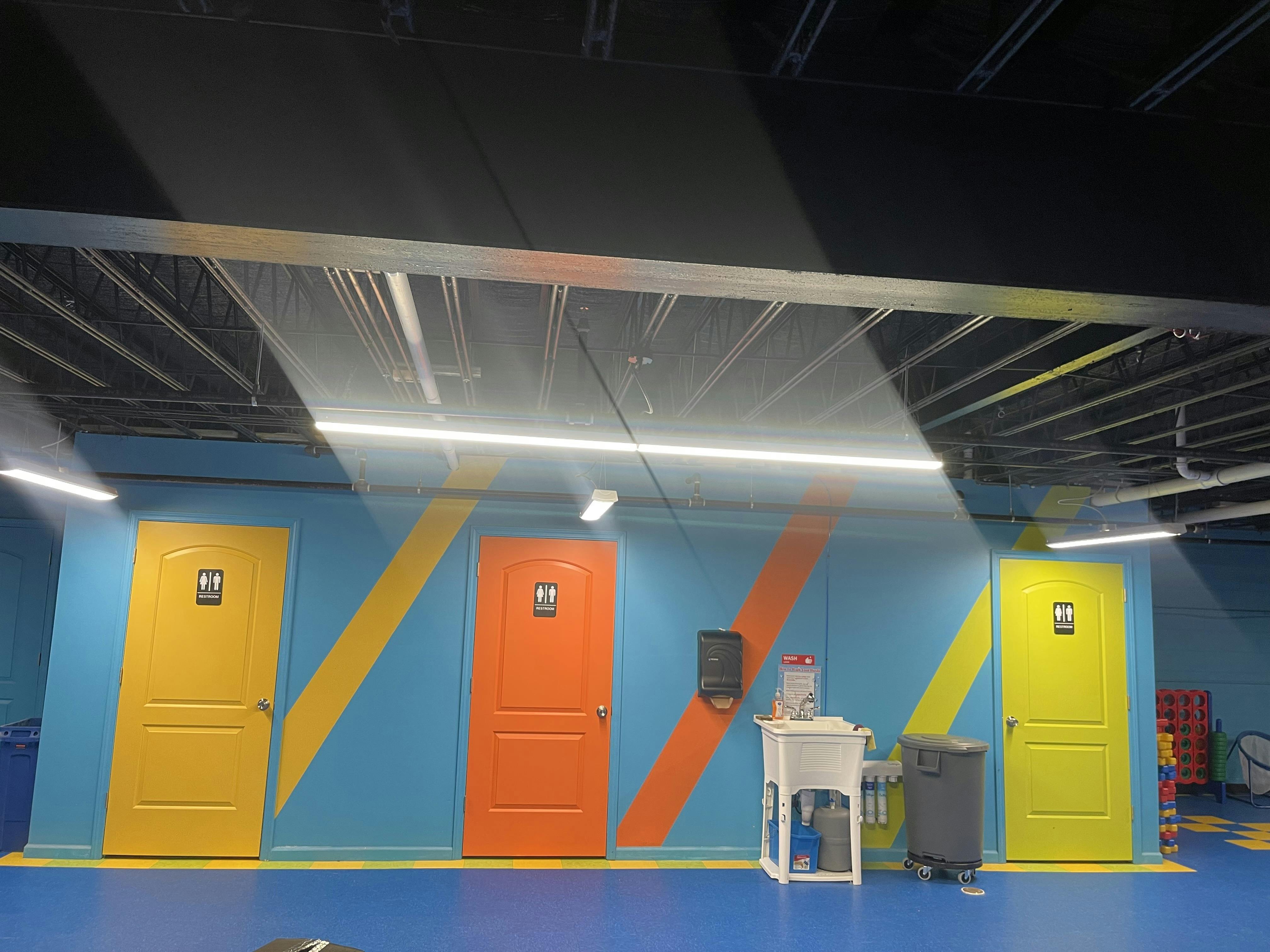Three colorful restroom doors with gender-neutral signs on a blue wall with stripes