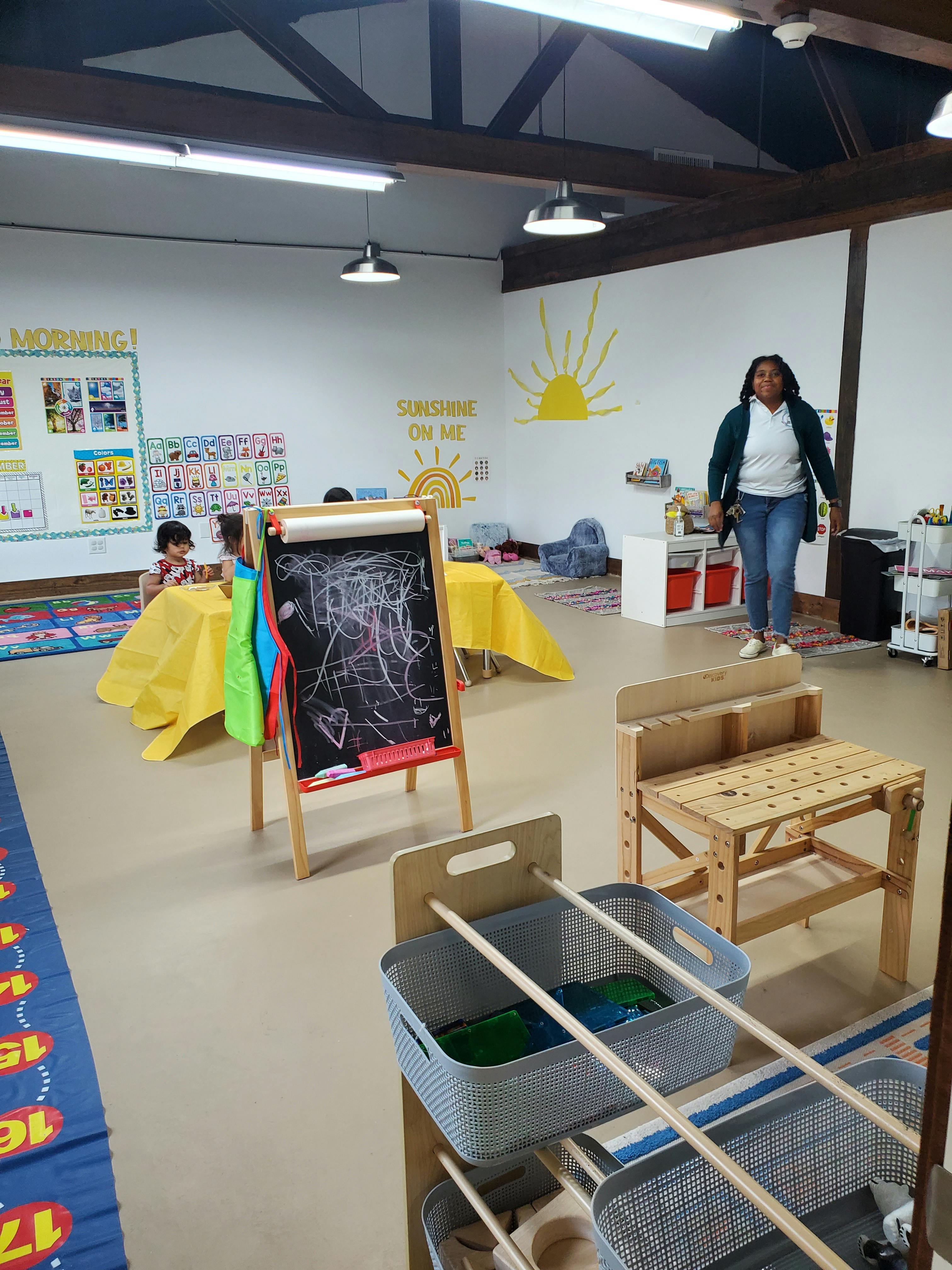 Person standing in a classroom with children at tables and an easel