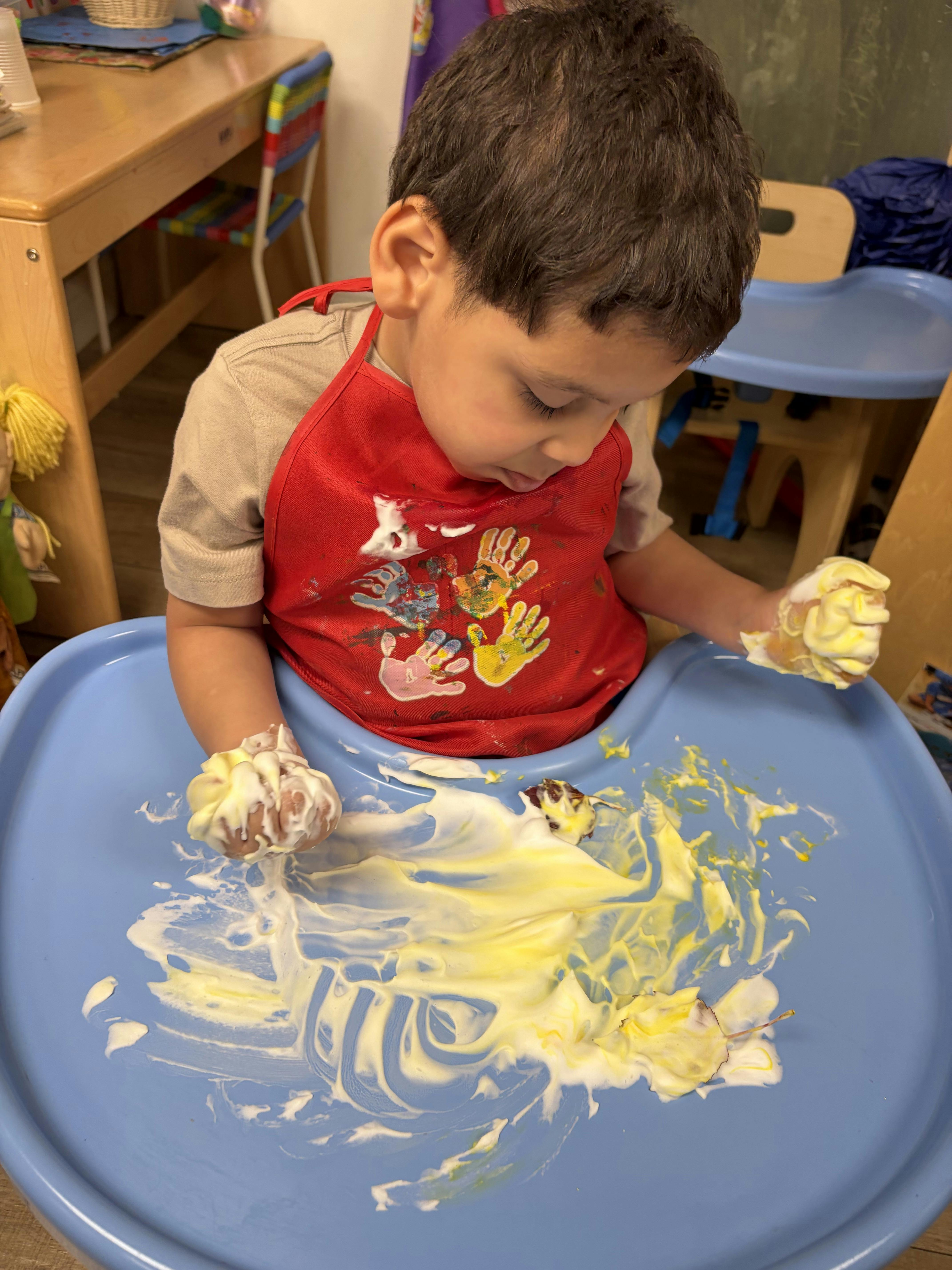Child playing with yellow paint and foam on a high chair tray